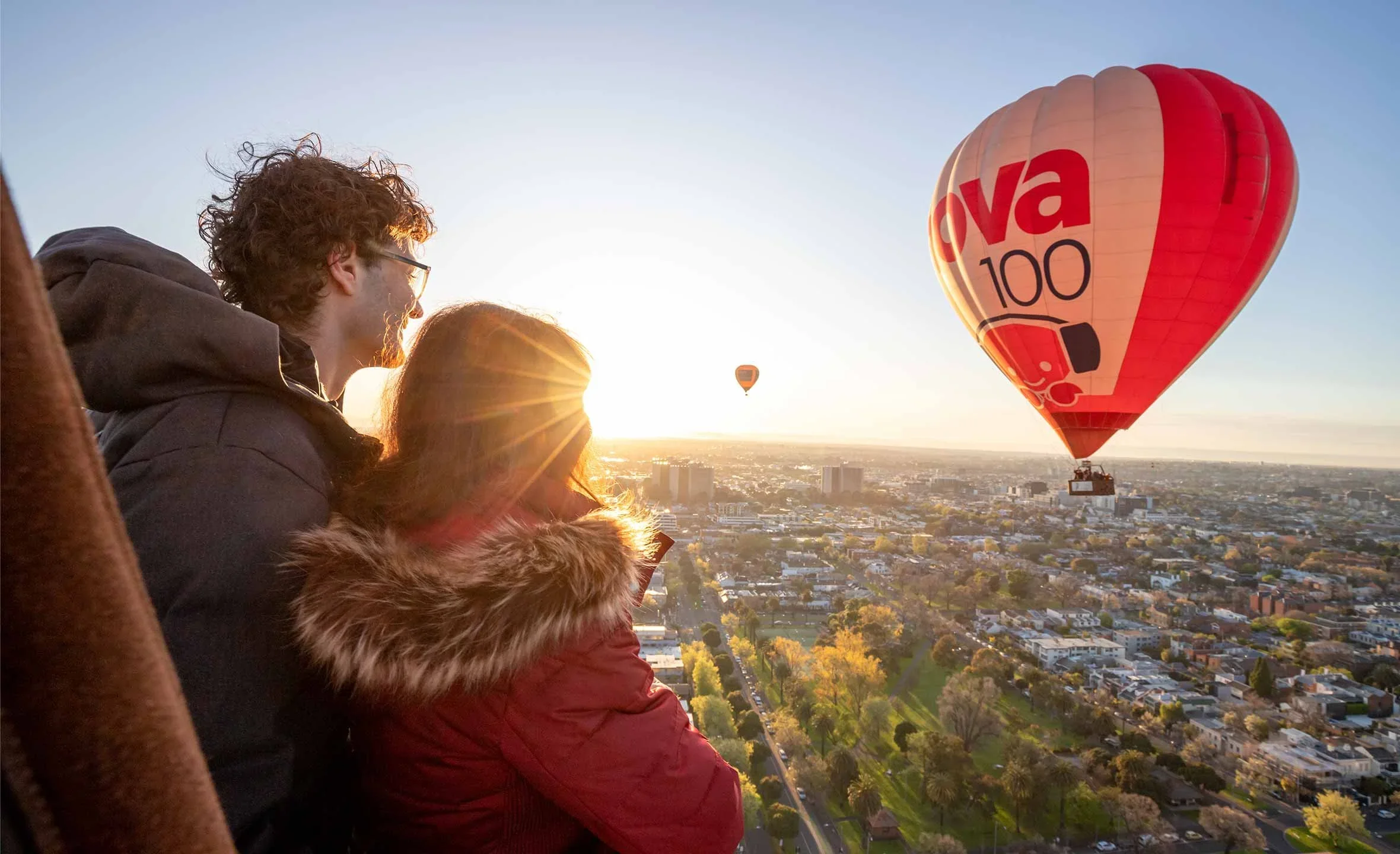 Two people in a hot air balloon watching sunset with cityscape and other hot air balloons in the background.