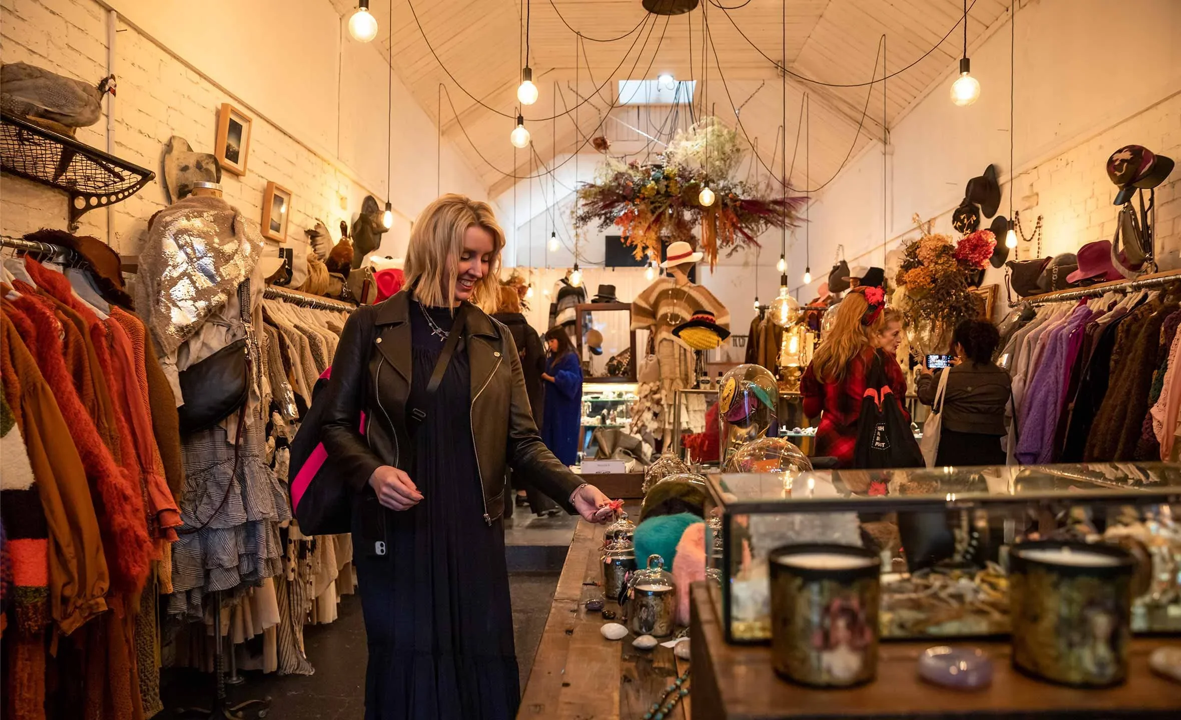 A woman shopping in a boutique with racks of colorful clothing, hats, and accessories, decorated with hanging light bulbs and floral arrangements.