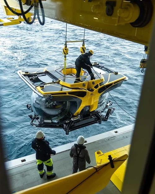 A yellow underwater exploration vehicle being lowered into the water from a large boat, with crew members in safety gear observing.