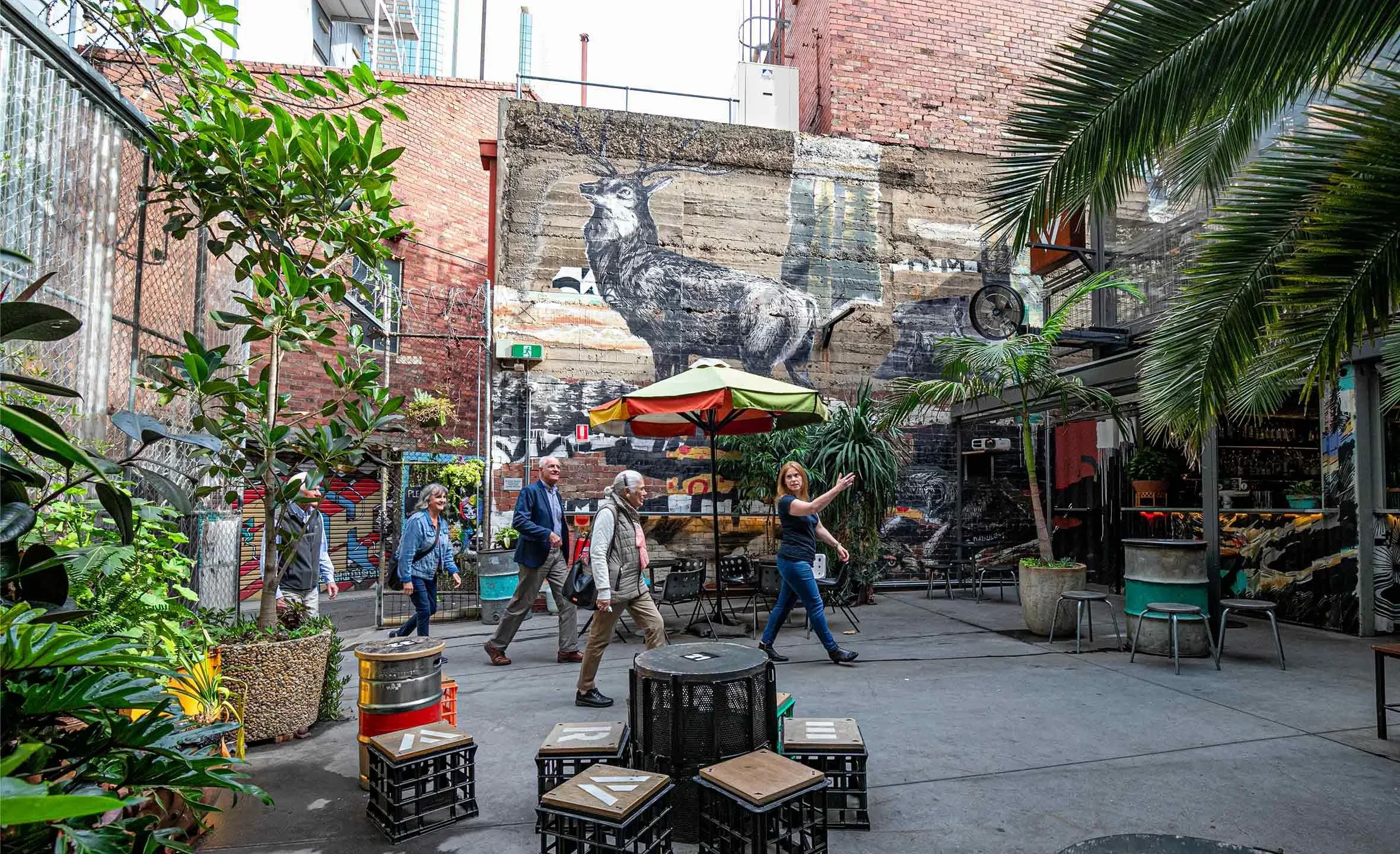 People walking through a vibrant outdoor urban courtyard with lush green plants, colorful graffiti murals, and a large mural of a deer on a brick wall in the background. There are tables, chairs, and a multicolored umbrella providing shade.