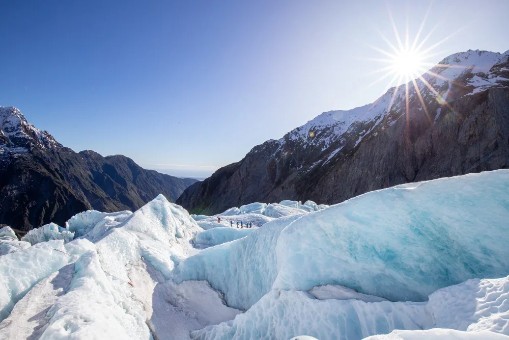 Mountain landscape with glaciers, snow, and ice, with a bright sun shining overhead.