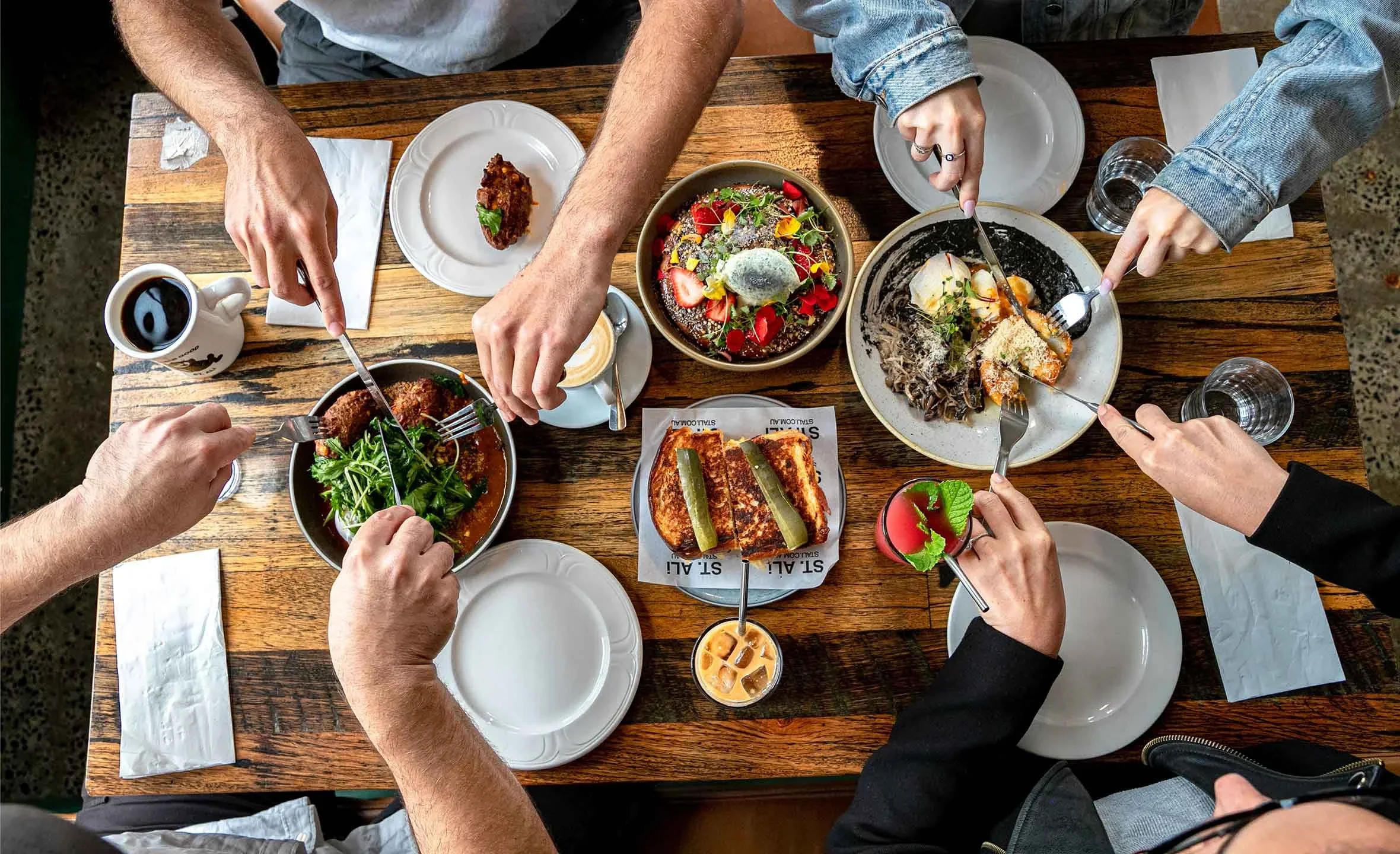 Overhead view of five people sharing a wooden table with various dishes, including salads, grilled items, a drink with ice, and a dessert. They are using forks and knives to serve themselves.