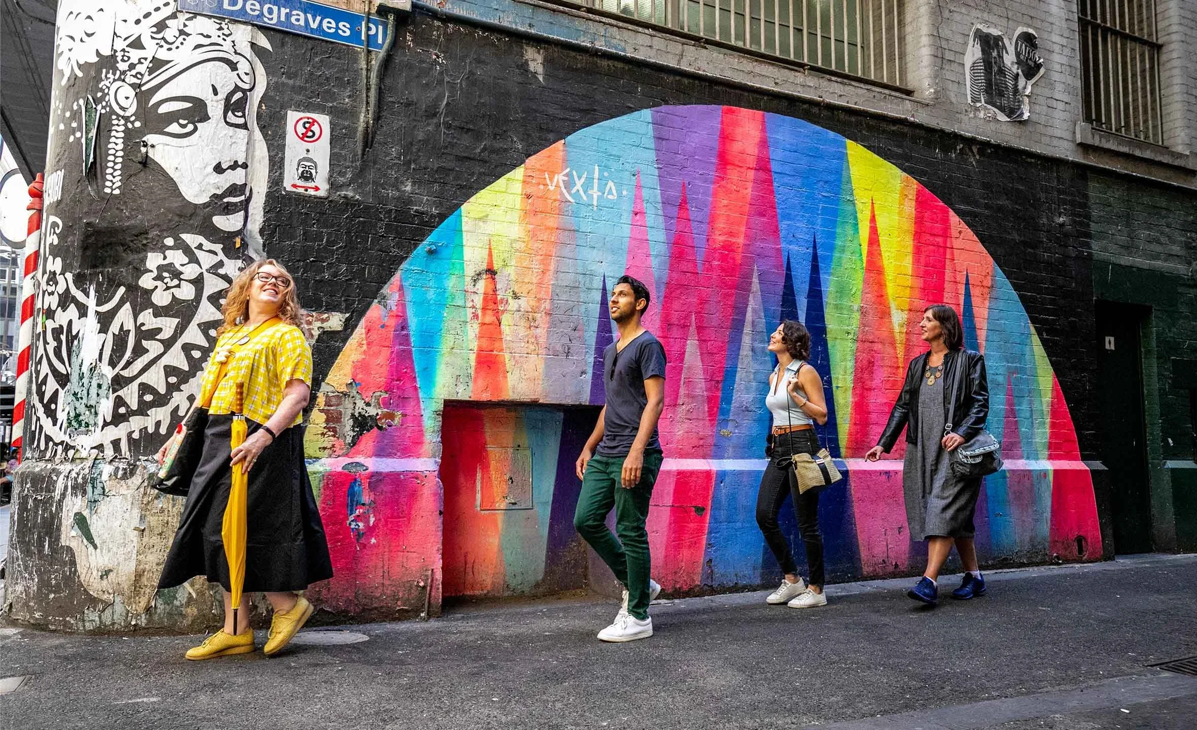 Four people walking past a graffiti mural on a city street wall. The mural features a large rainbow-colored circular design and a black-and-white portrait of a woman with flowers and jewelry. There are street signs and windows on the building.