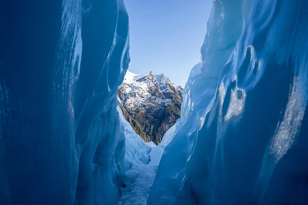 View through a narrow passage between large blue glaciers, with a snow-capped mountain in the background under a clear blue sky.