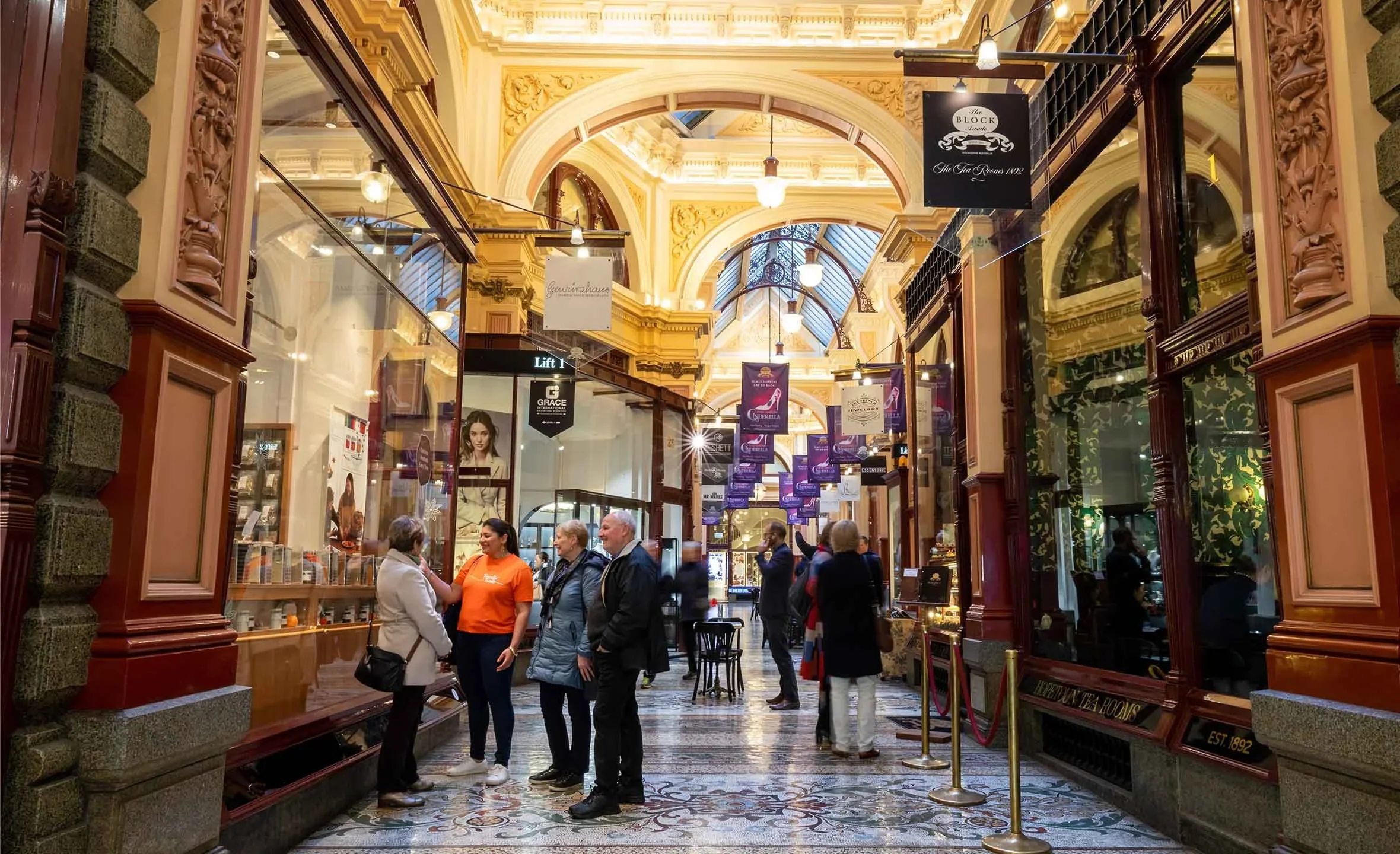 Inside a grand historic shopping arcade with ornate architectural details, multiple people are shopping and socializing, with shops on each side and decorative hanging banners.