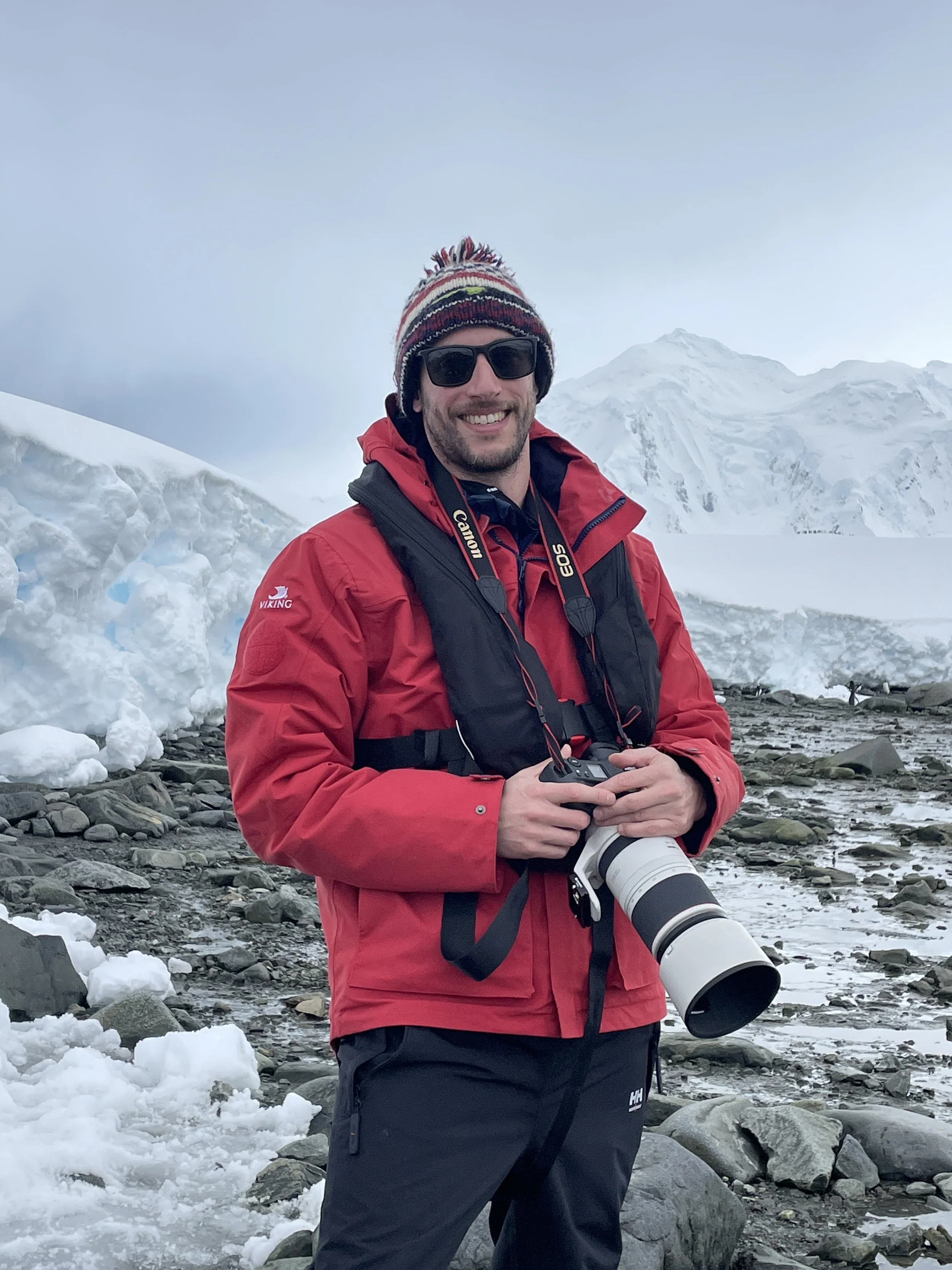 Justin Meneguzzi wearing a red jacket and striped beanie smiling while holding a camera, standing on rocky icy terrain in Antarctica with snow and glaciers in the background.