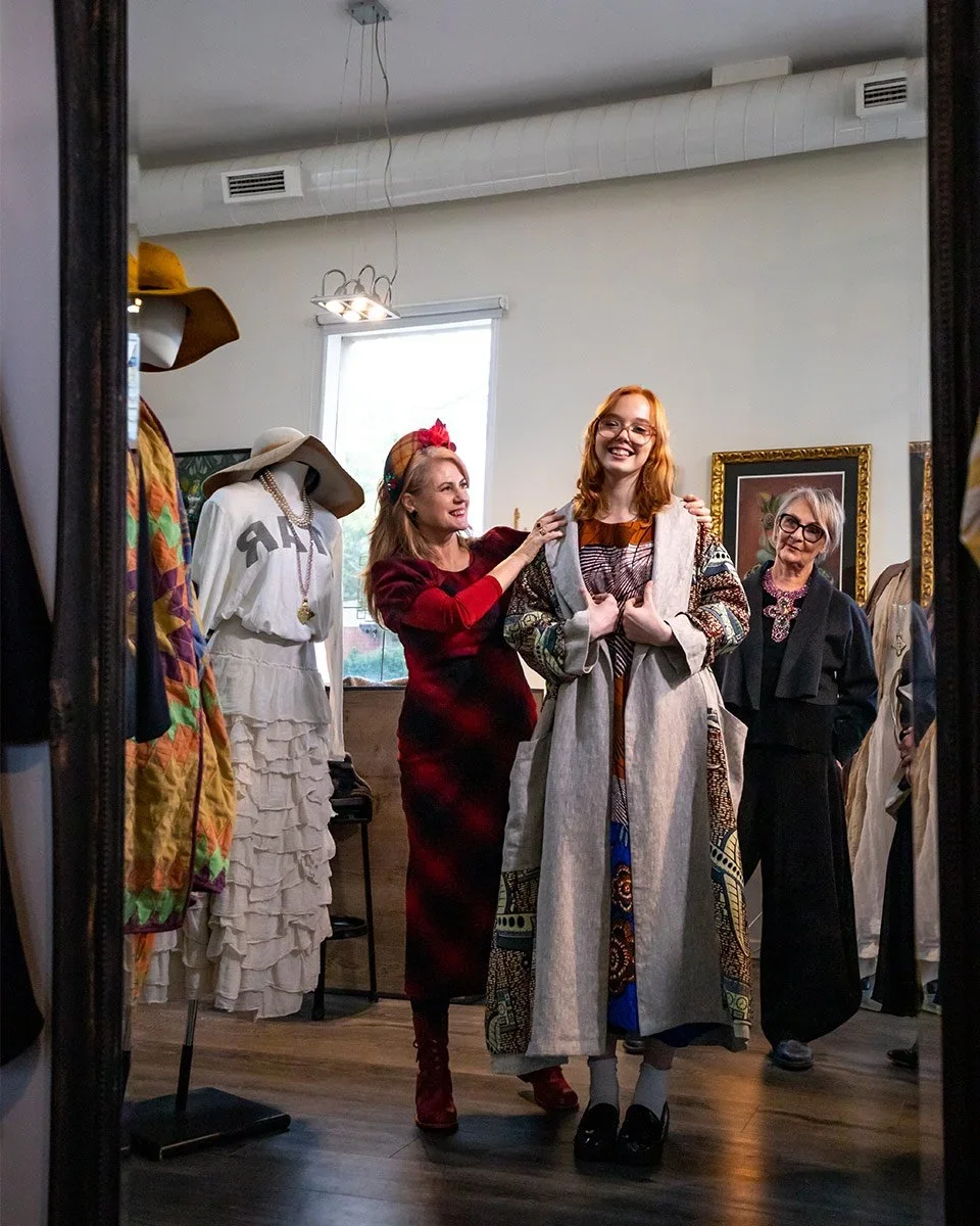 A woman in a red and black checkered dress trying on a colorful, patterned coat while being assisted by another woman in a red dress in a boutique. Two other women stand in the background, one with gray hair and glasses, observing and smiling. The bo