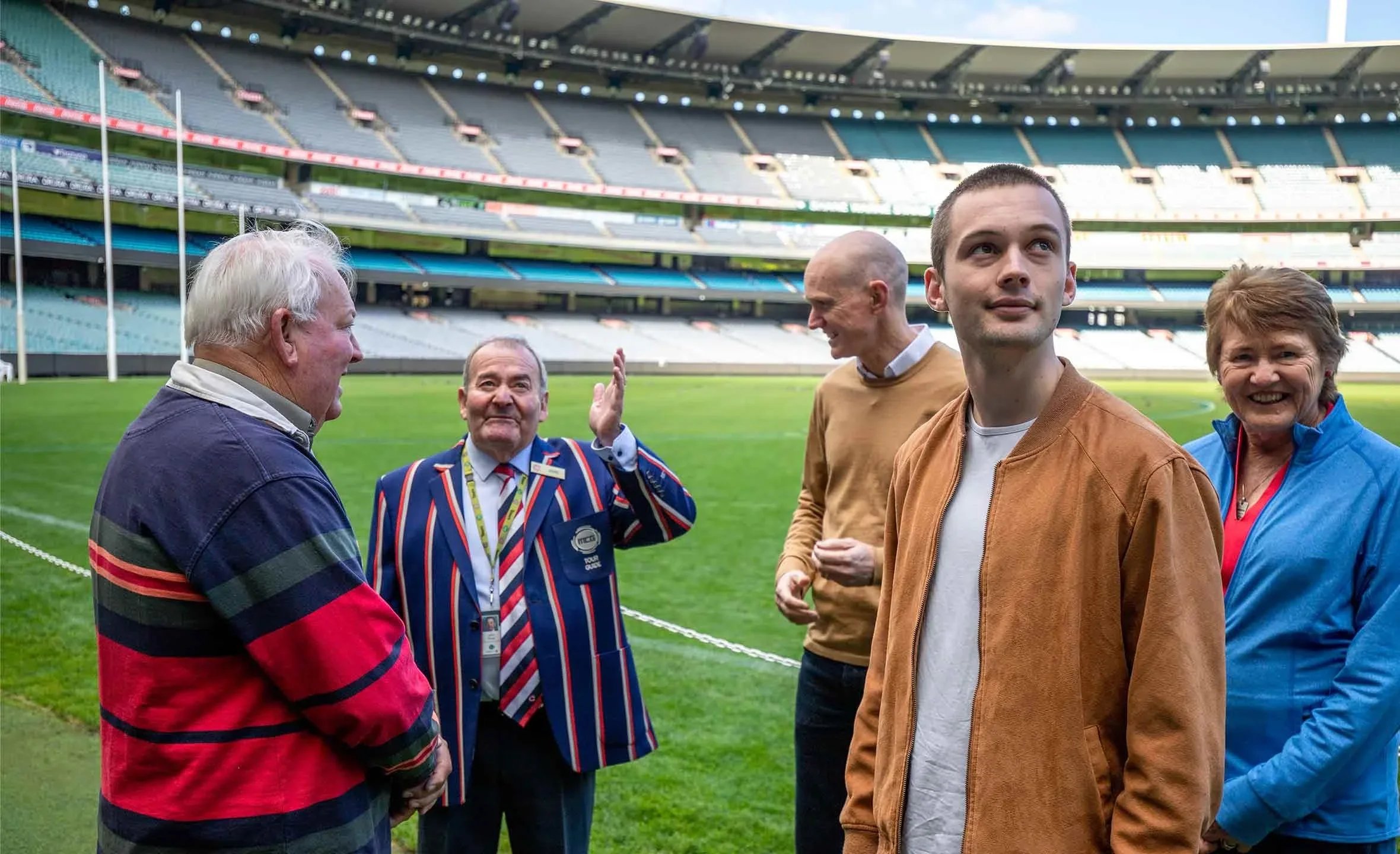 A group of five people standing on a sports field inside a stadium, engaging in conversation and smiling, with the empty stadium seats visible in the background.