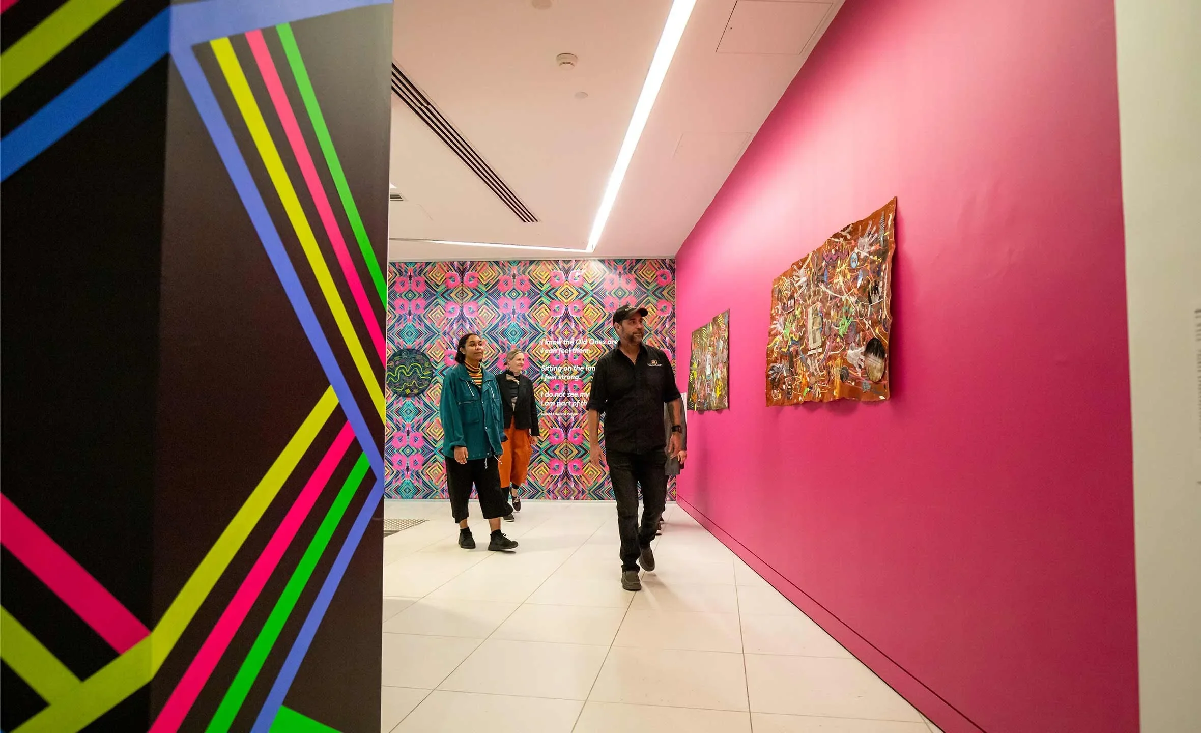 Visitors in an art gallery with colorful and vibrant artwork, including a large pink wall with painted pieces, and a background with geometric patterns in pink, green, and blue.