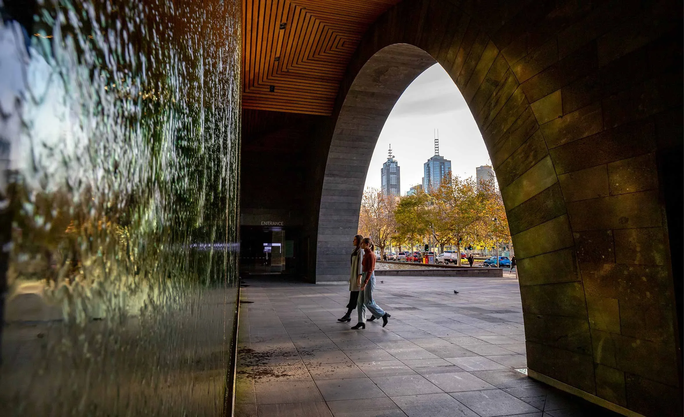 Two women walking through an archway under a modern cityscape with tall buildings and autumn trees.