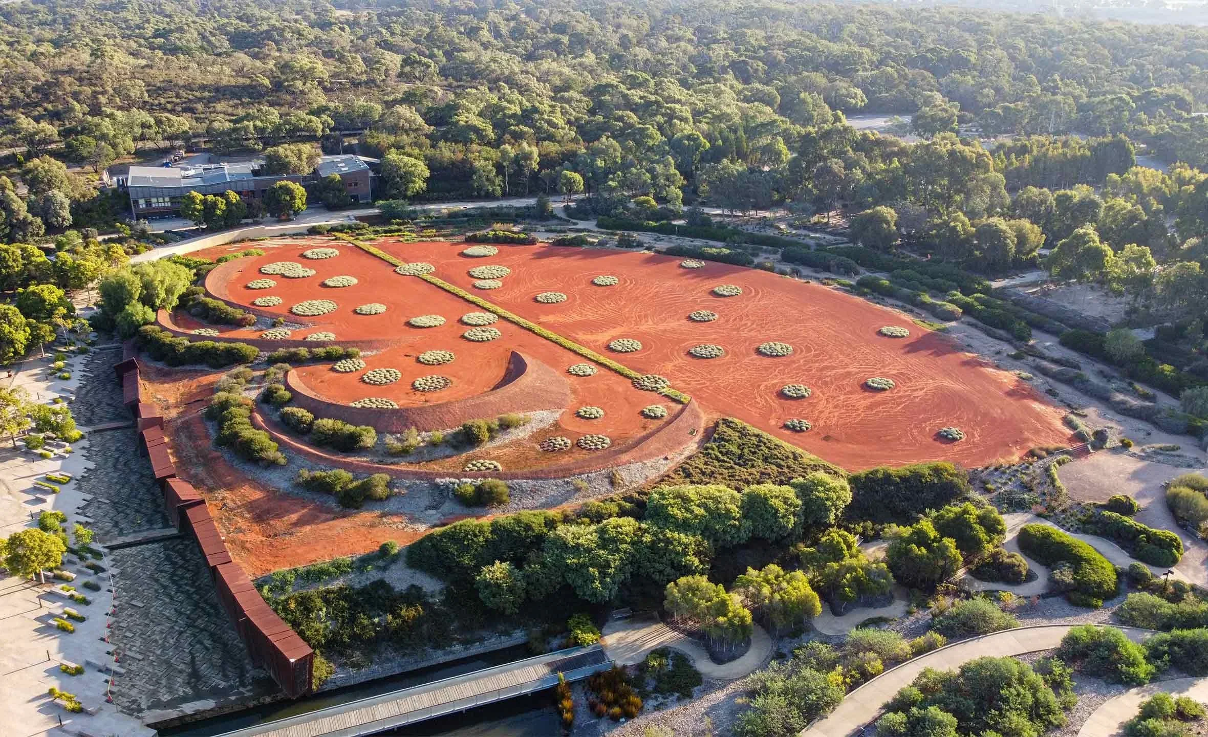 An aerial view of a landscaped park with red sandy areas, circular plant arrangements, greenery, trees, walking paths, and surrounding buildings.