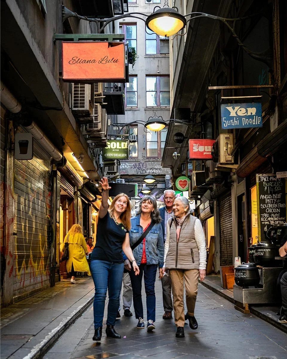 Four people are walking and talking down a narrow, urban alleyway with various shop signs and graffiti. The group appears to be enjoying their time together.