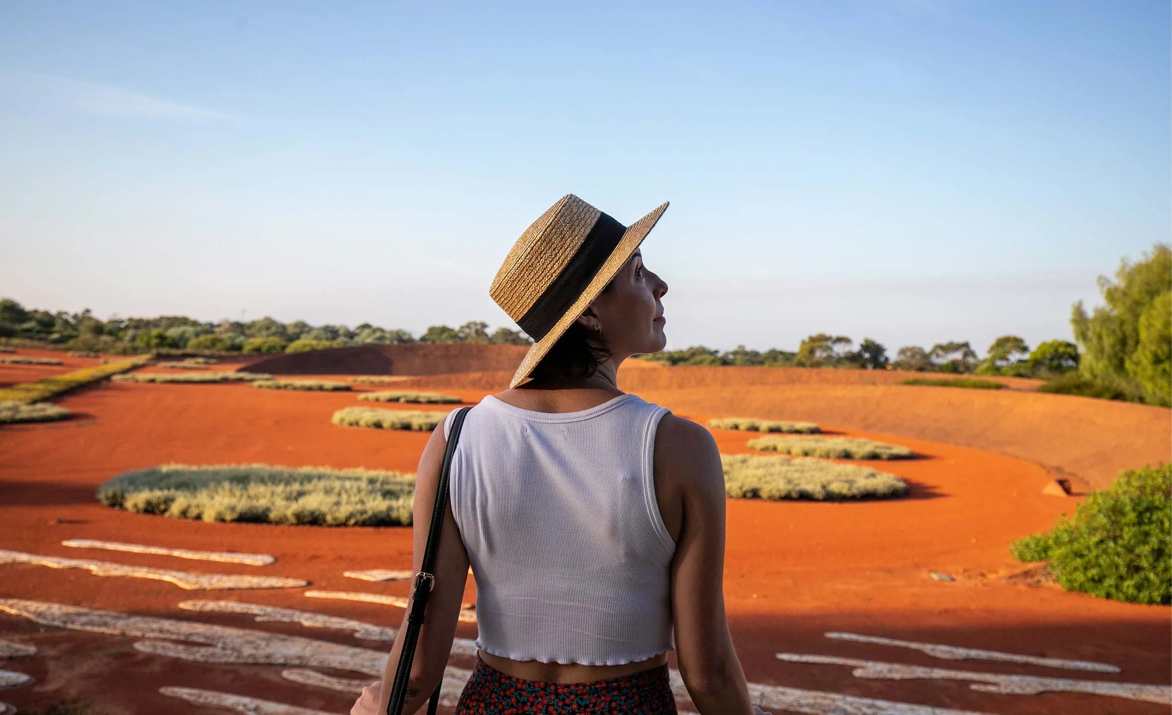 Woman wearing a straw hat and sleeveless top standing outdoors on a red hill with sparse vegetation, looking up at the sky during daytime.