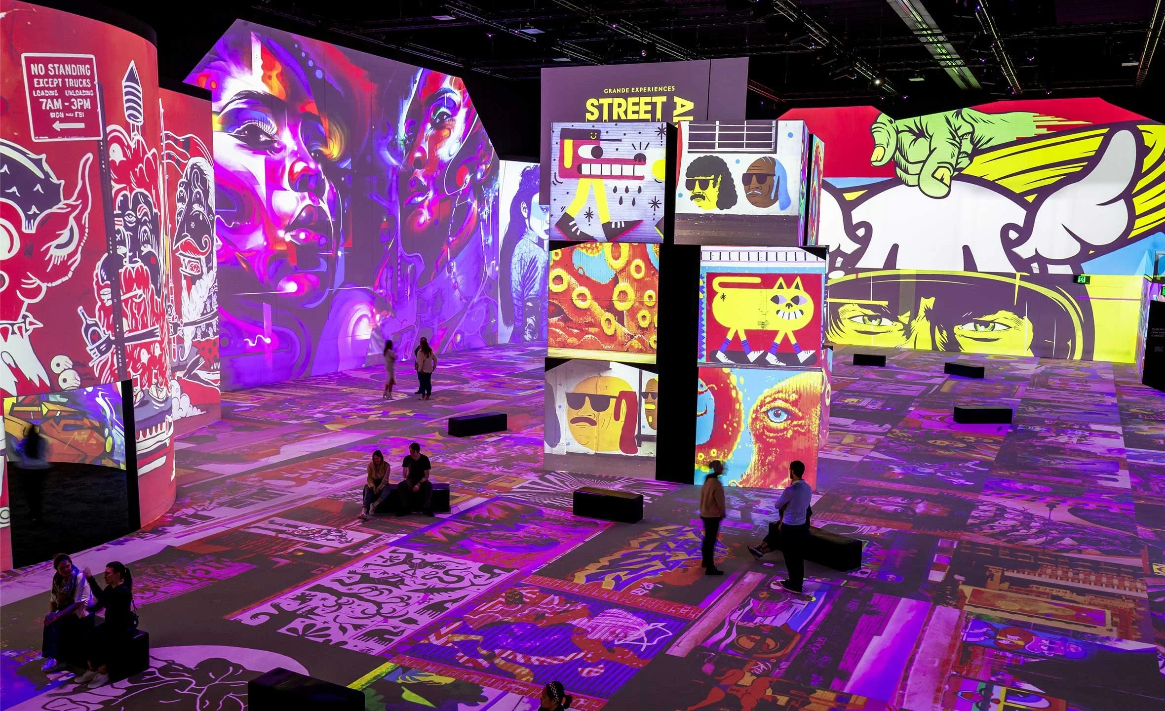 Colorful digital art installation with large screens displaying graffiti-style and pop art images, with visitors exploring the space.