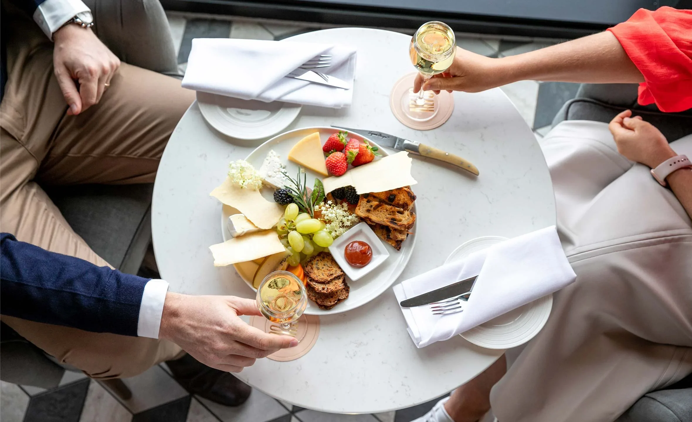A round marble table with a cheese platter, strawberries, grapes, crackers, and jam, surrounded by three people holding glasses of wine, with white napkins and utensils on the table.