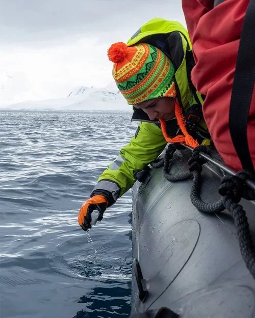 Person in colorful winter hat and green jacket leaning over the side of a boat, examining the water near the boat's edge in a cold, snowy environment.