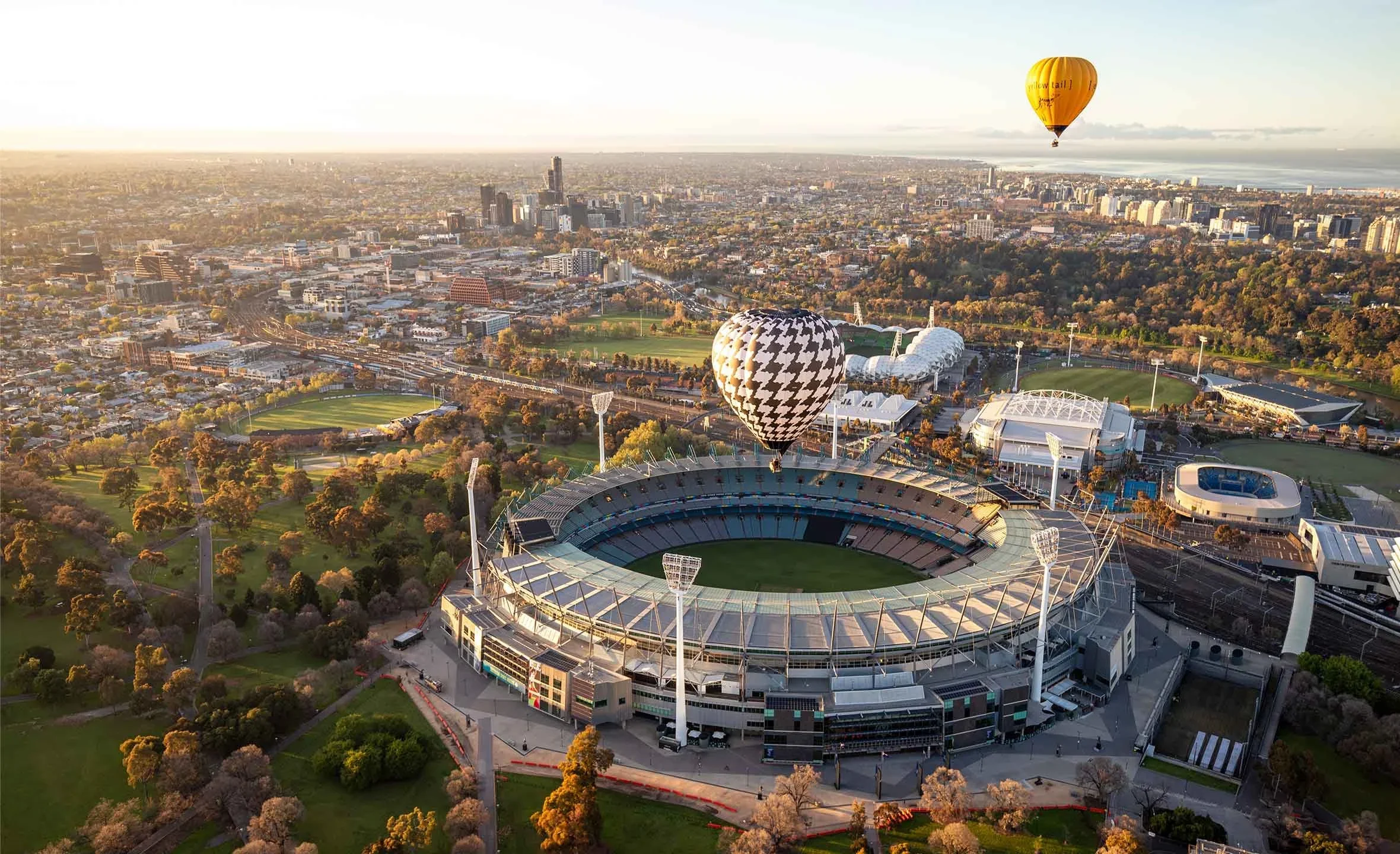 Aerial view of a sports stadium with houndstooth and yellow hot air balloons floating above, city skyline in the background, during sunset.