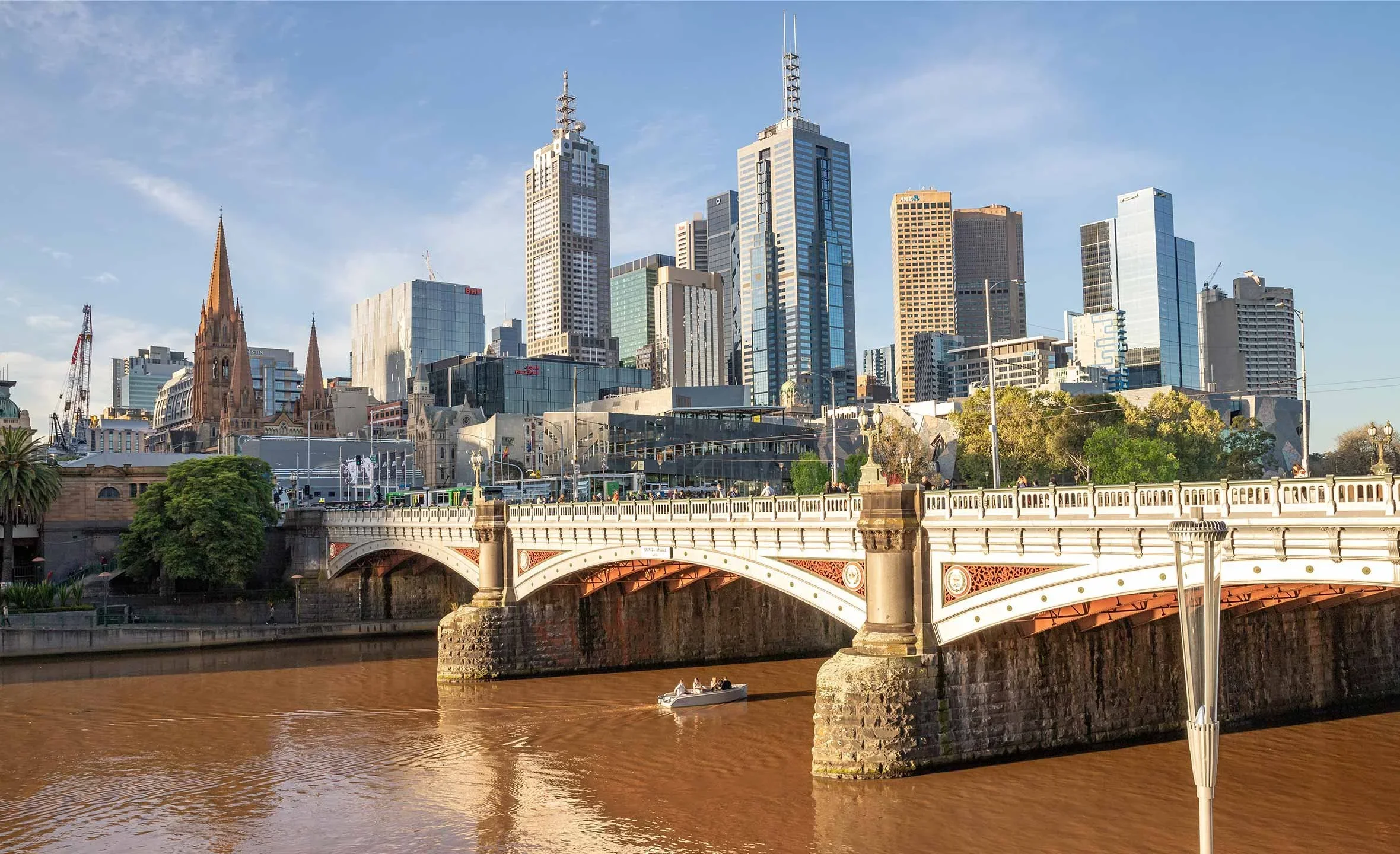 Melbourne skyline with tall modern skyscrapers, historic church with pointed steeples, bridge over river with a boat, and clear blue sky.