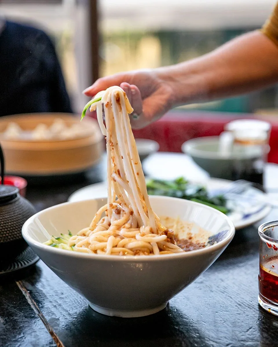 Person lifting noodles from a white bowl of Asian-style noodles with sauce, on a dining table with condiments and side dishes.