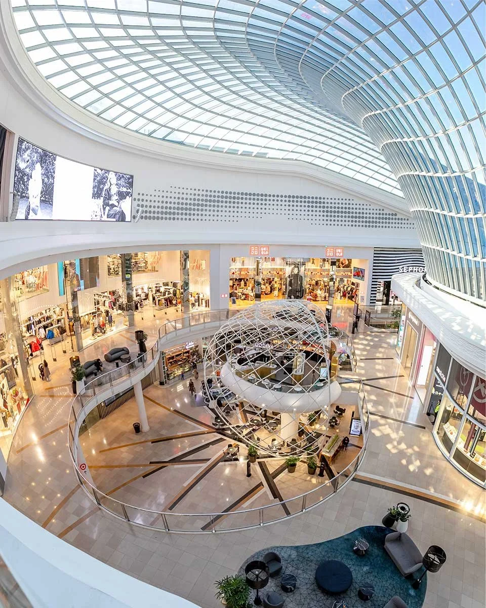 Interior view of a modern shopping mall with a large glass ceiling, retail stores, and a circular sitting area with a globe structure in the center.