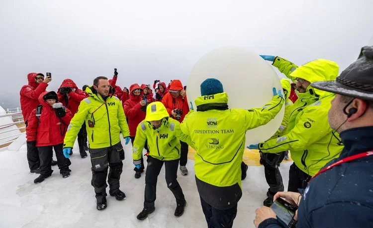 A group of expedition team members wearing bright yellow jackets working together to move a large snow or ice block, with onlookers in red jackets taking photos in a snowy environment.