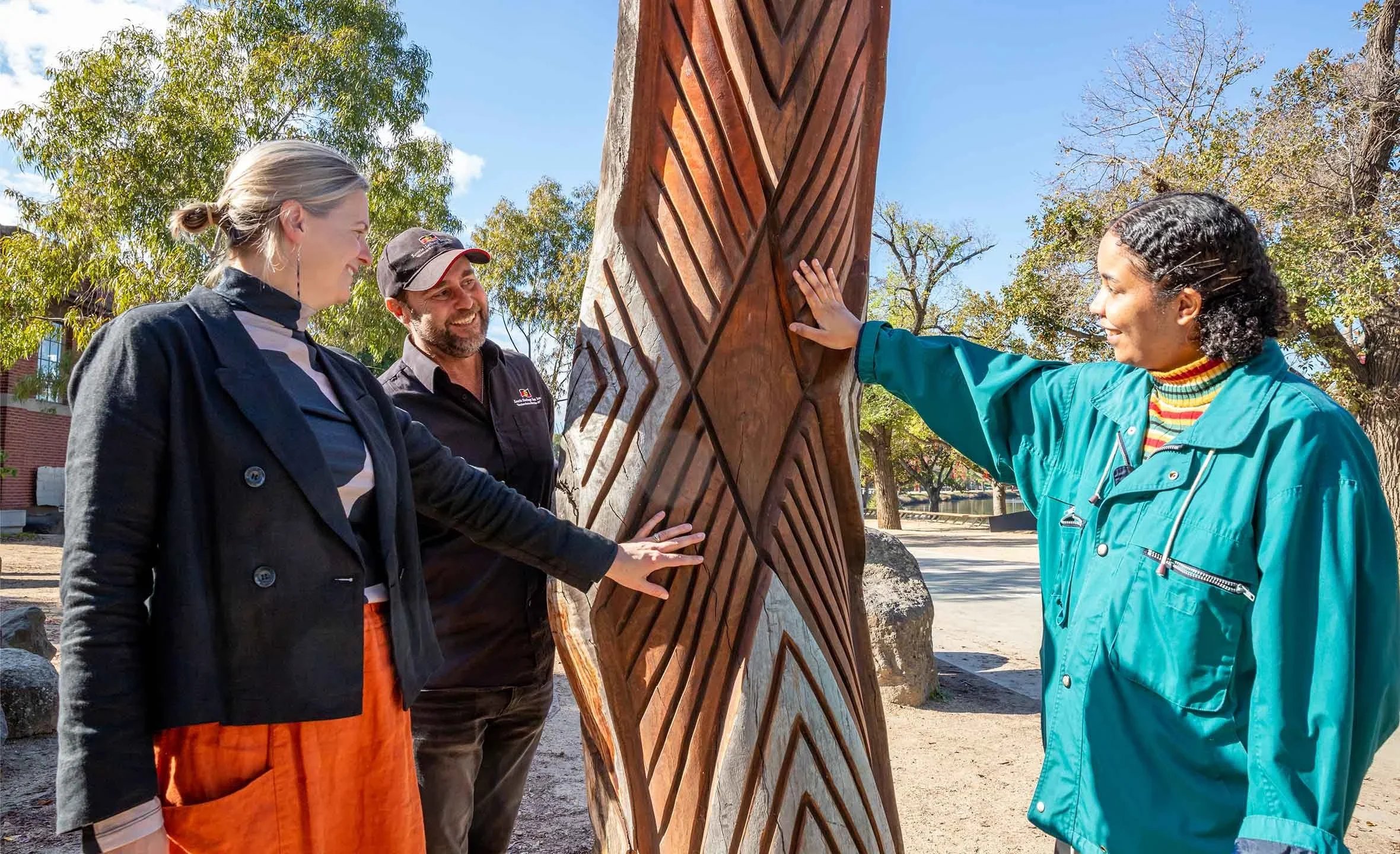 Three people outdoors around a carved wooden sculpture, with trees and a building in the background. One woman in a teal jacket touches the sculpture, smiling, while a man and another woman observe and smile.