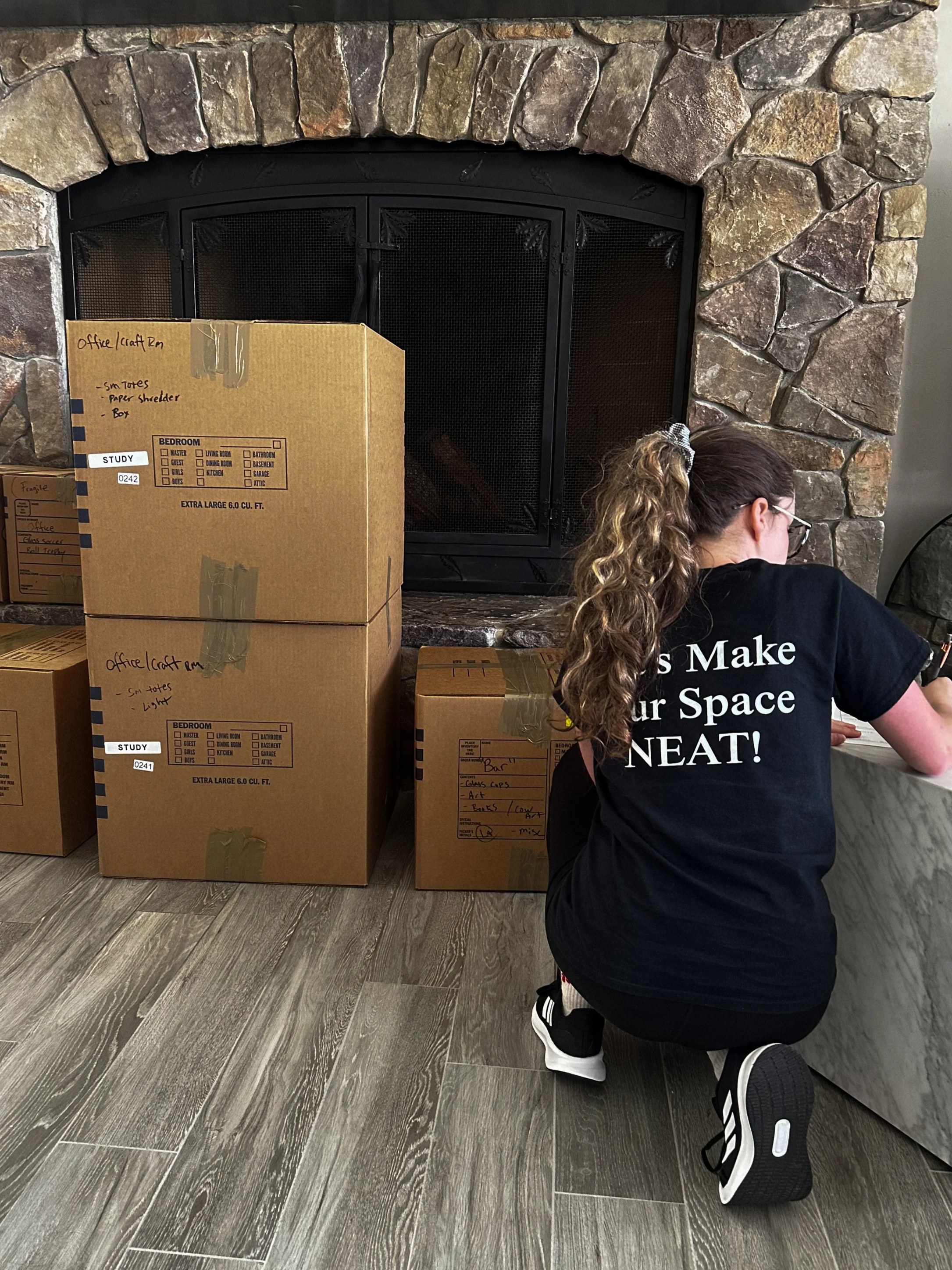A room with hardwood floor, large window with curtains, a chandelier on the ceiling, a decorative mirror leaning against the wall, and several cardboard boxes labeled 'Bedroom,' 'Kitchen,' and 'Fragile'.