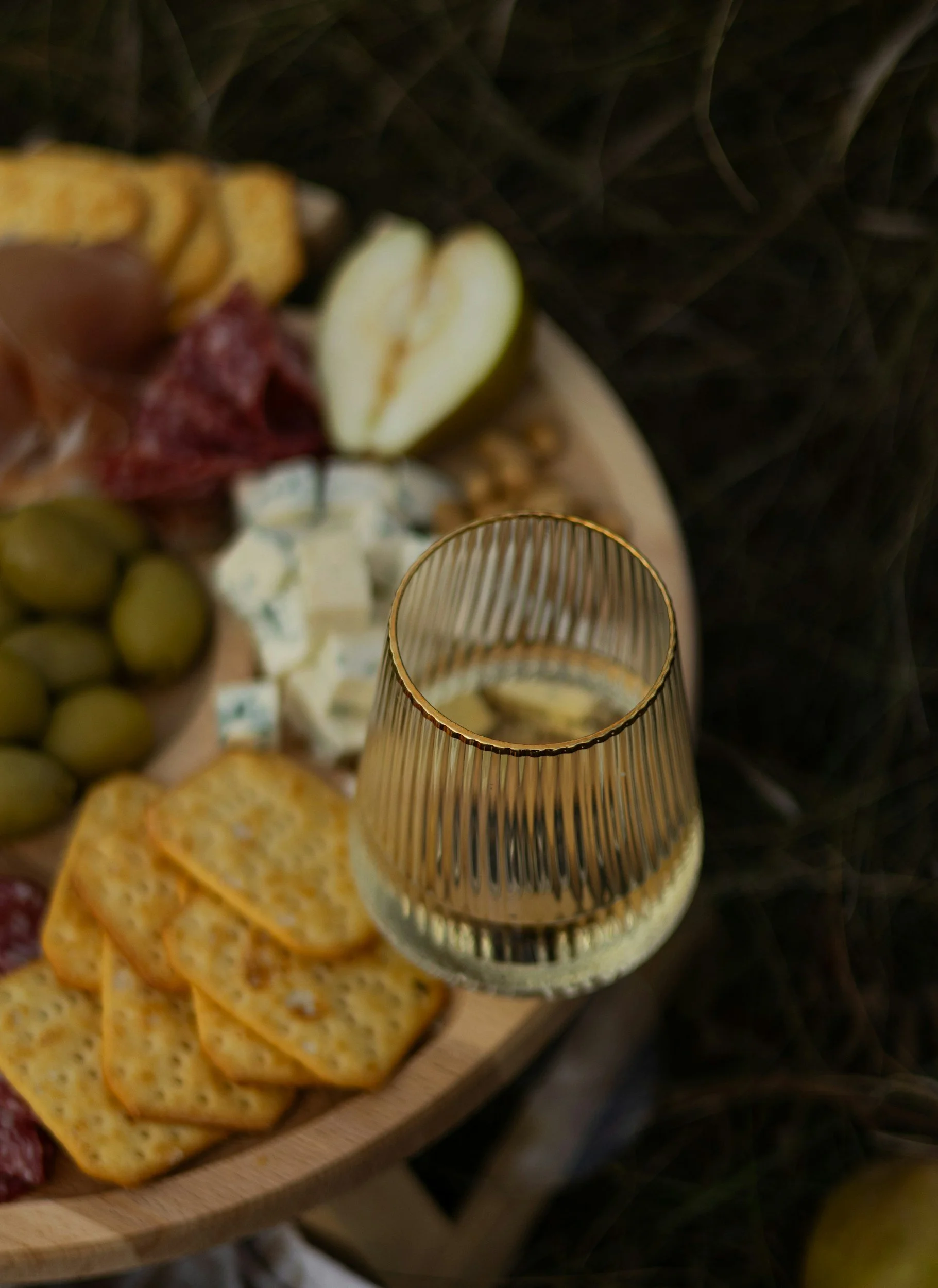 Close-up of a glass of white wine on a wooden platter with assorted cheese, crackers, grapes, pear slices, cured meats, and blue cheese, set against a dark background.