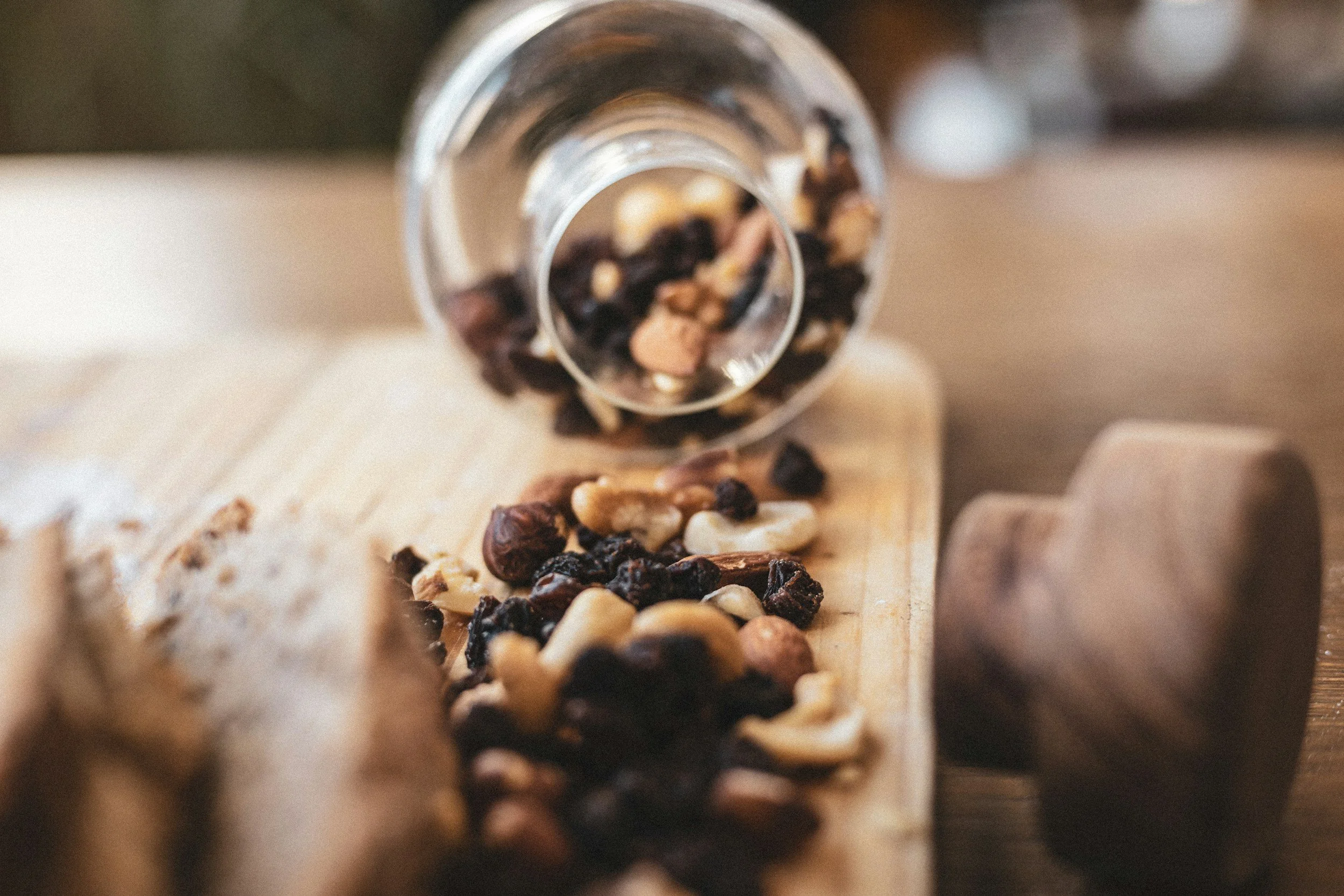 Mixed dried fruits and nuts spilling from a glass container onto a wooden cutting board.