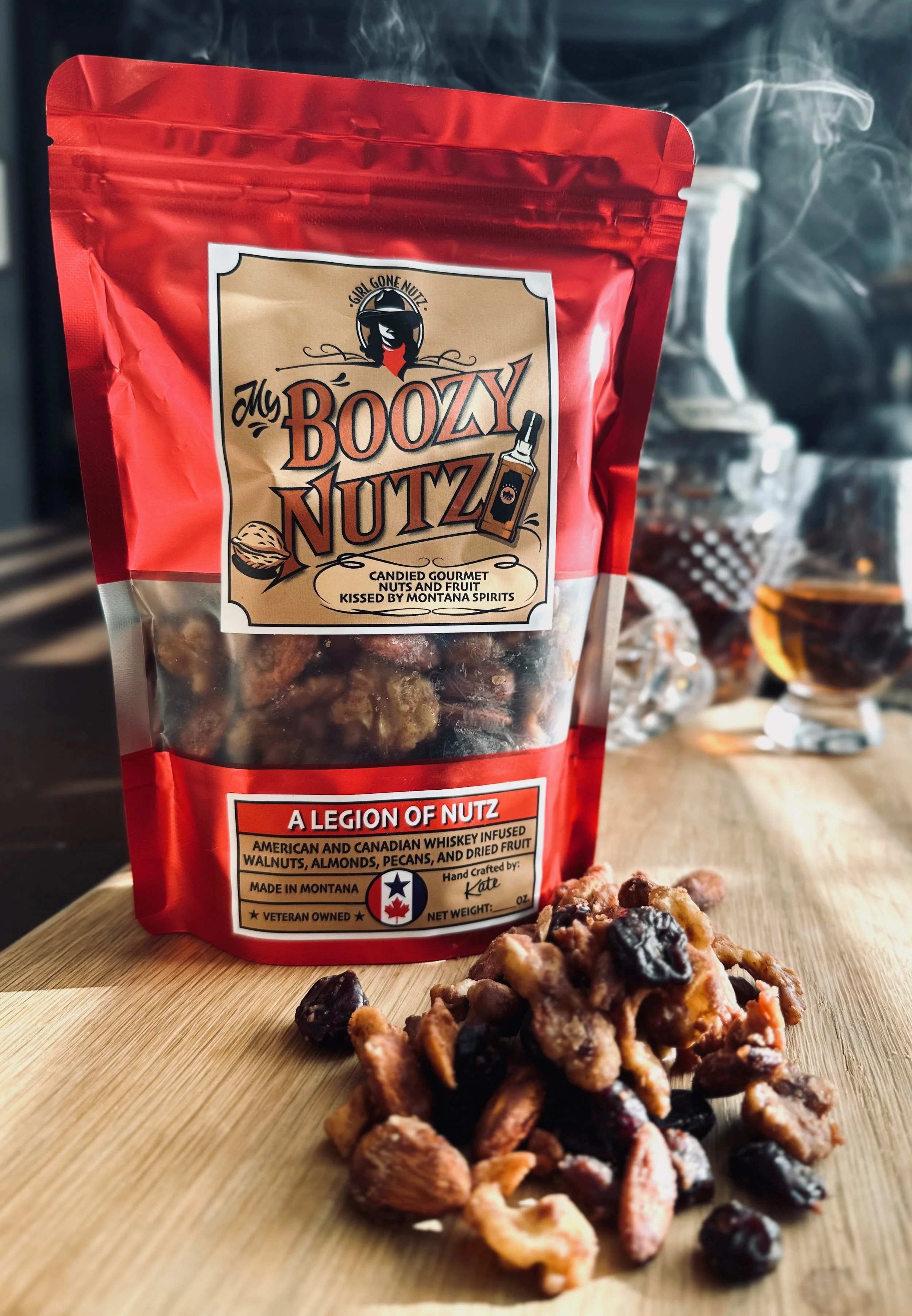 A red bag of Girl Gone Nutz's My Boozy Nutz cashew and dried fruit mix on a wooden surface, with a pile of the mix in front and a glass of dark beverage in the background.