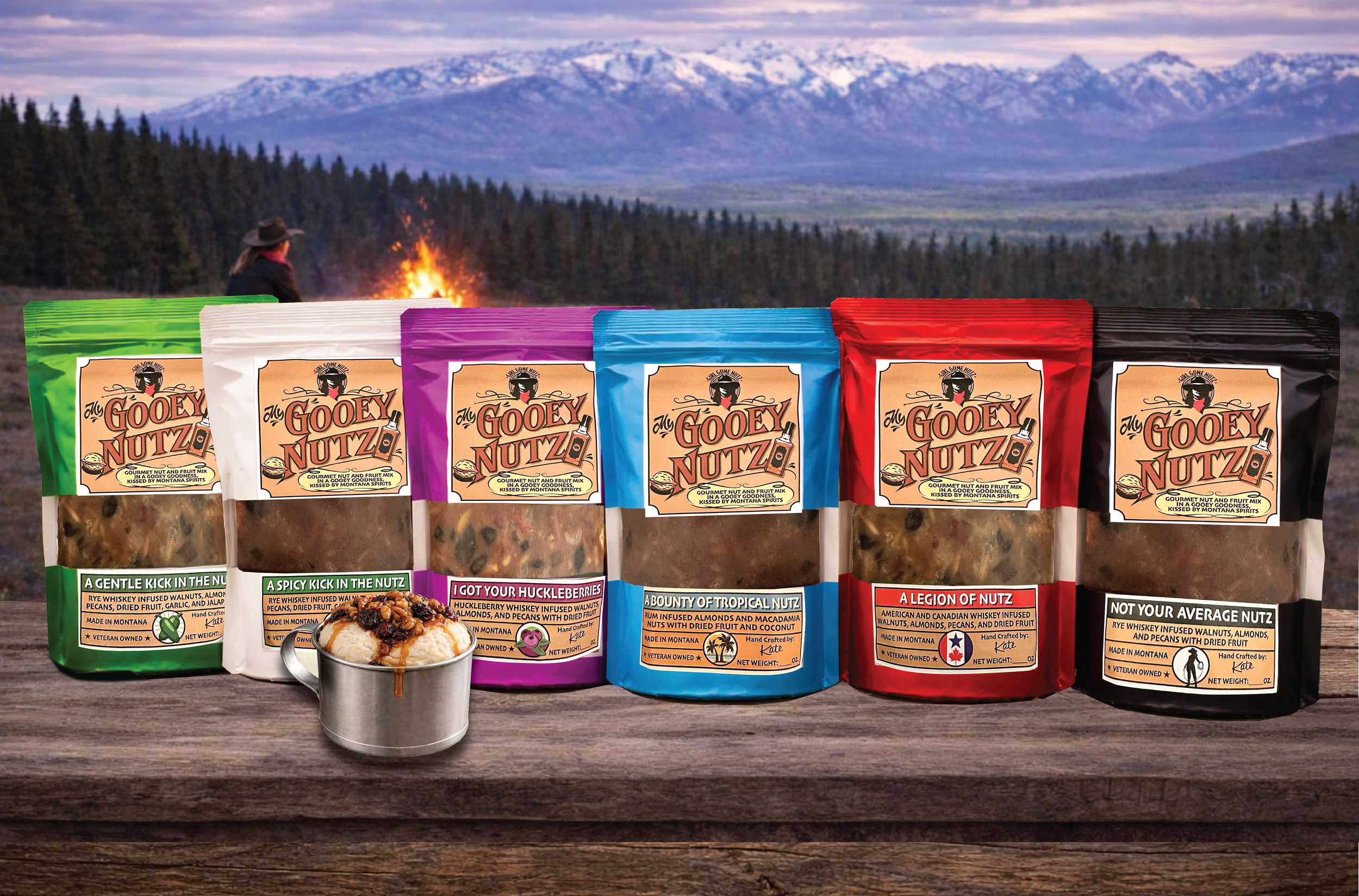 Six bags of GOOEY NUTZ snack mix in green, white, purple, blue, red, and black packaging, with a mug of caramel sauce in front, on a wooden surface outdoors with a mountain landscape and a campfire in the background.