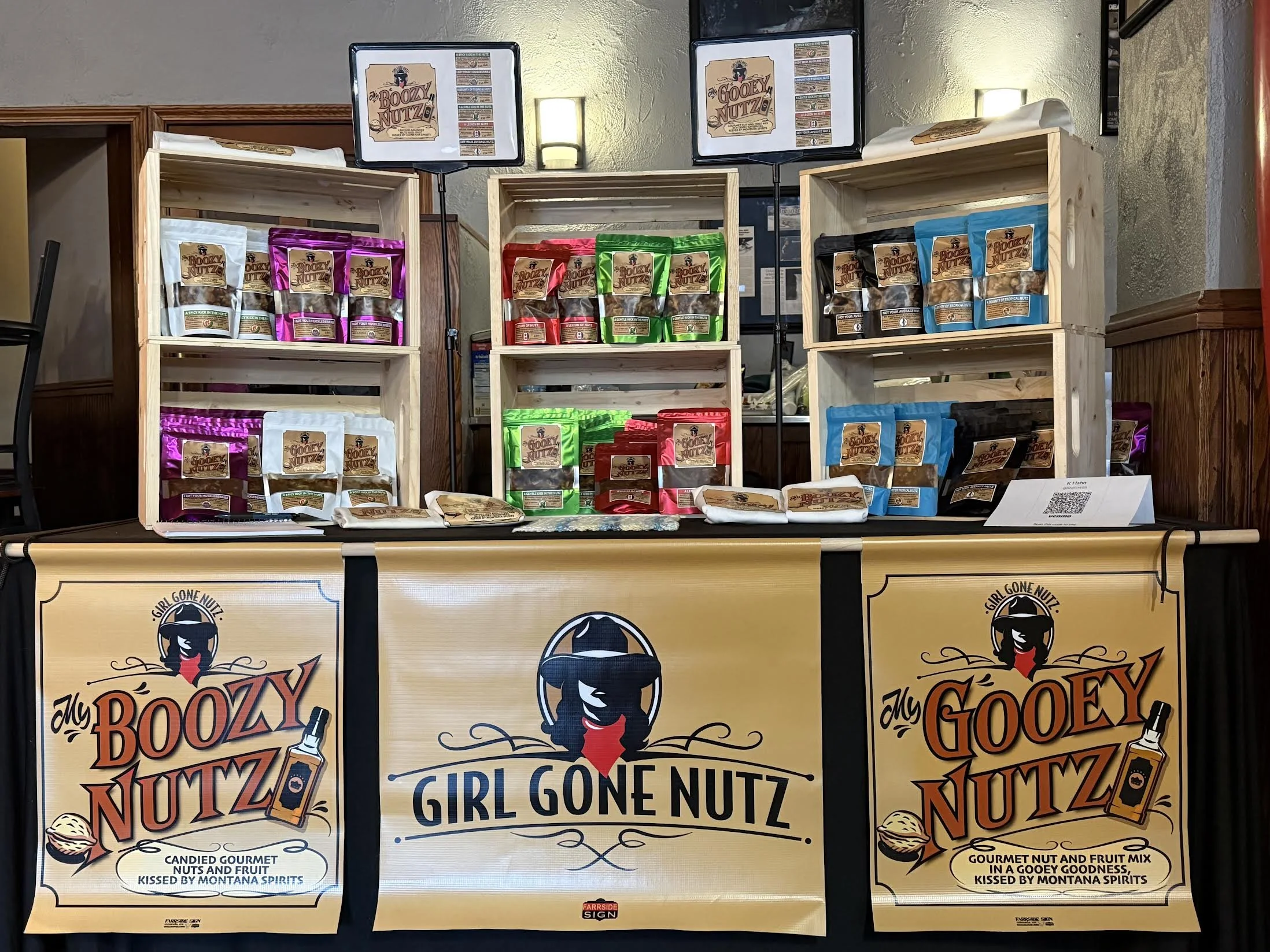Display of colorful packaged nut and fruit products on wooden shelves with promotional signs, including banners for 'My Boozy Nutz' and 'Girl Gone Nuttz' at a market or event.