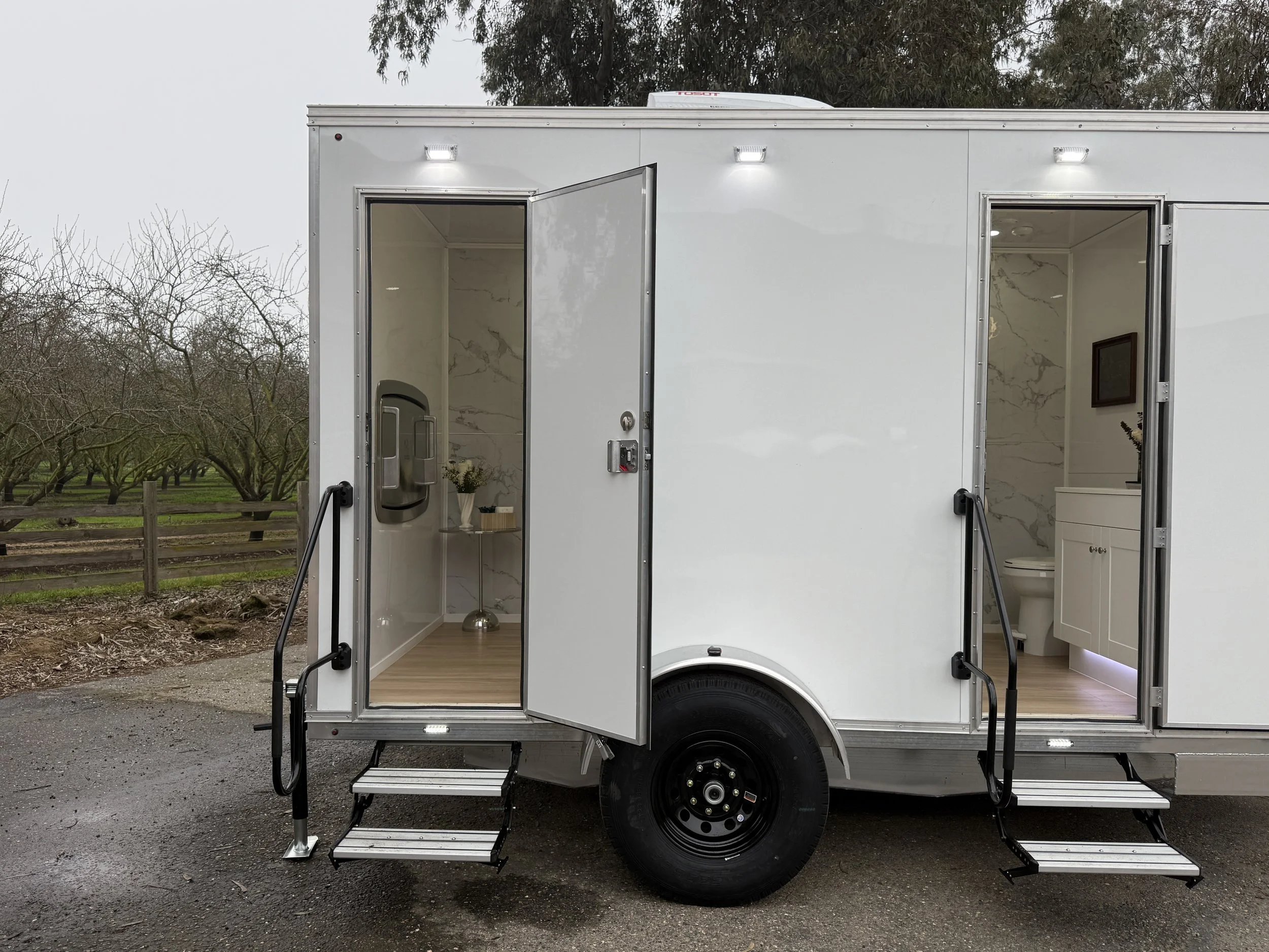Interior views of a mobile restroom trailer with two open doors, showing a sink on the left and a toilet on the right, set outdoors on a cloudy day.