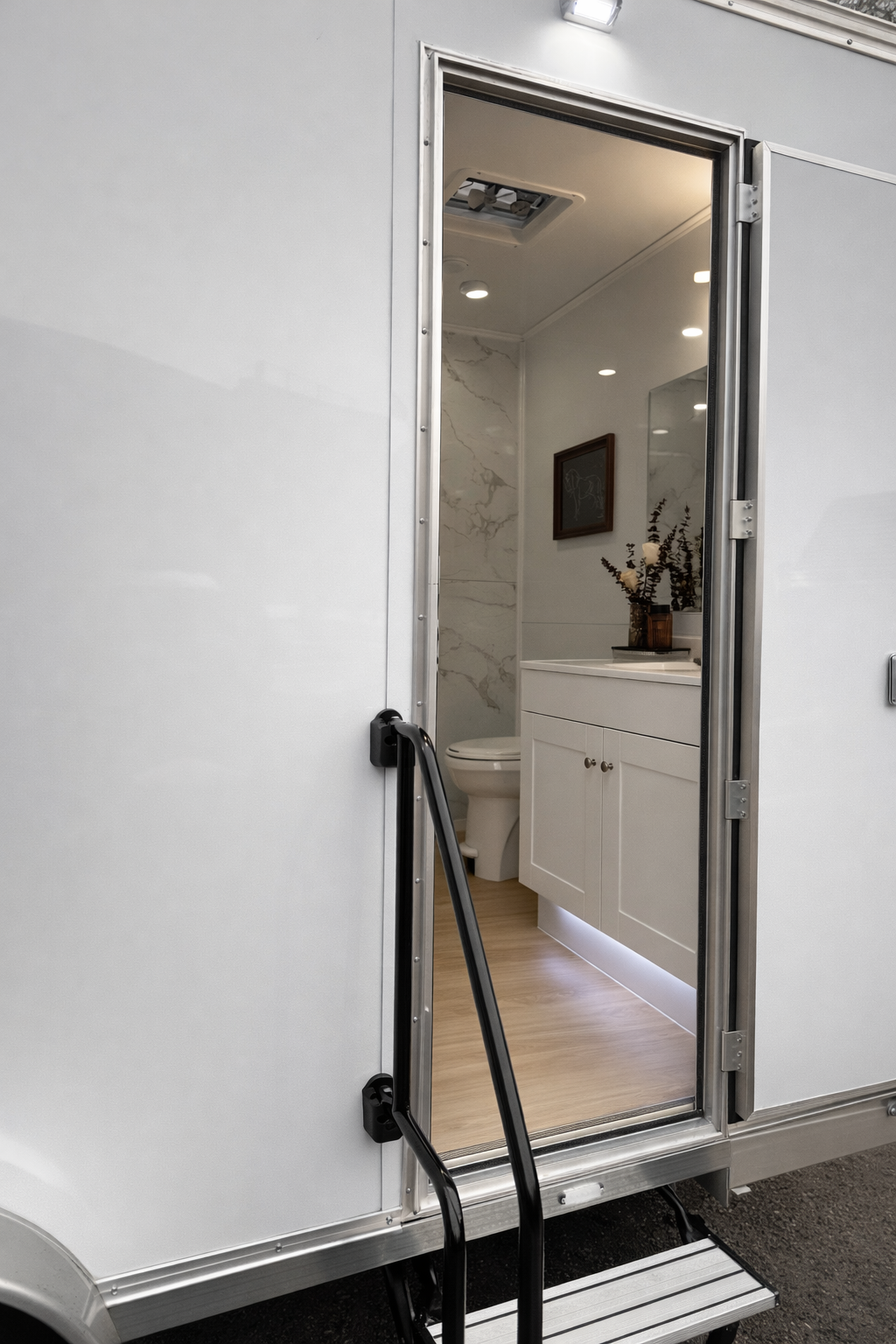 Exterior view of a tiny home or trailer with an open door revealing a modern bathroom inside, including a toilet, vanity with a sink, and wall decorations.