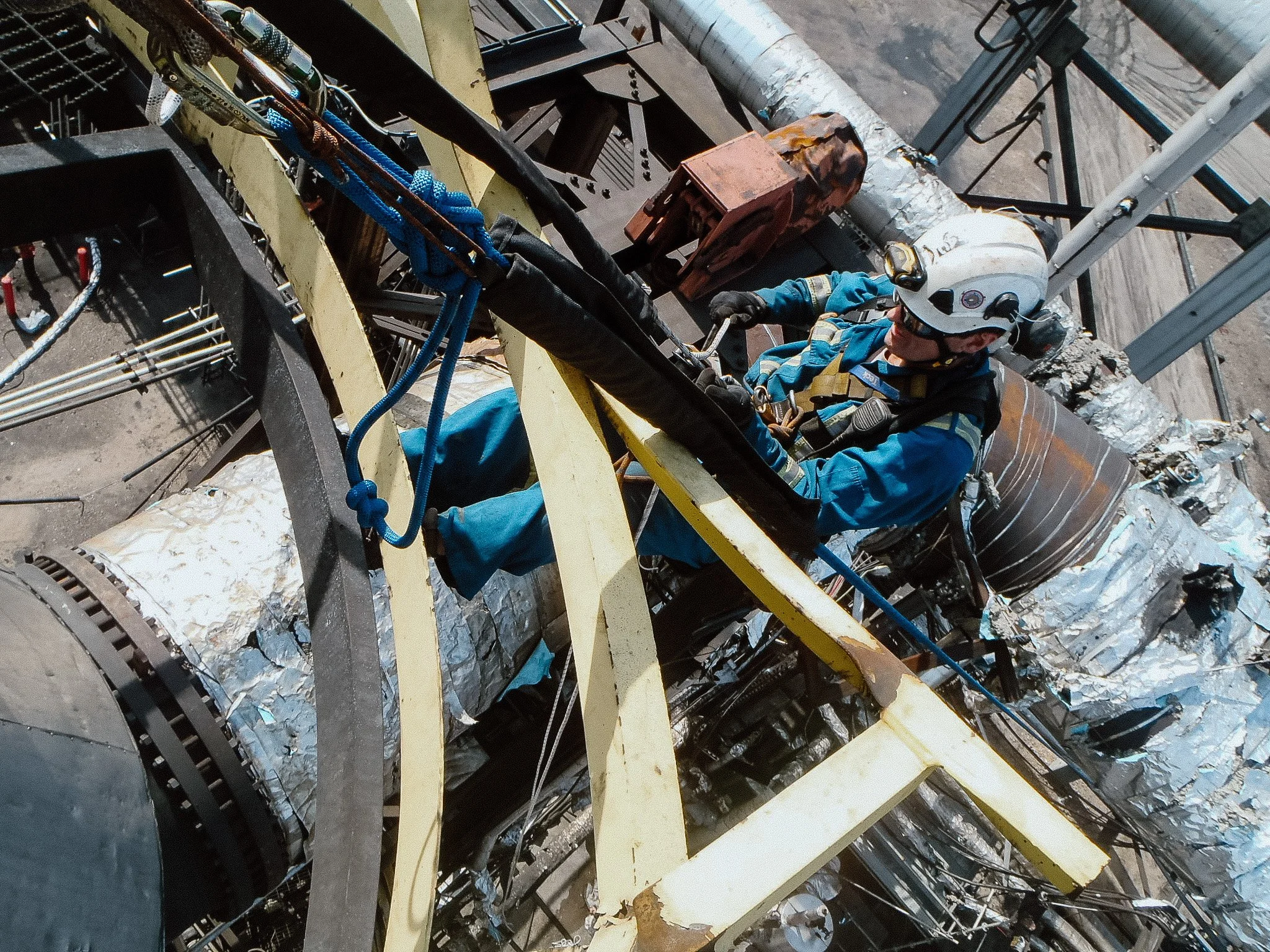Bruce Walker, founder of Creston Electric, wearing blue protective clothing, helmet, and harness working on a high industrial structure with metal pipes and machinery.