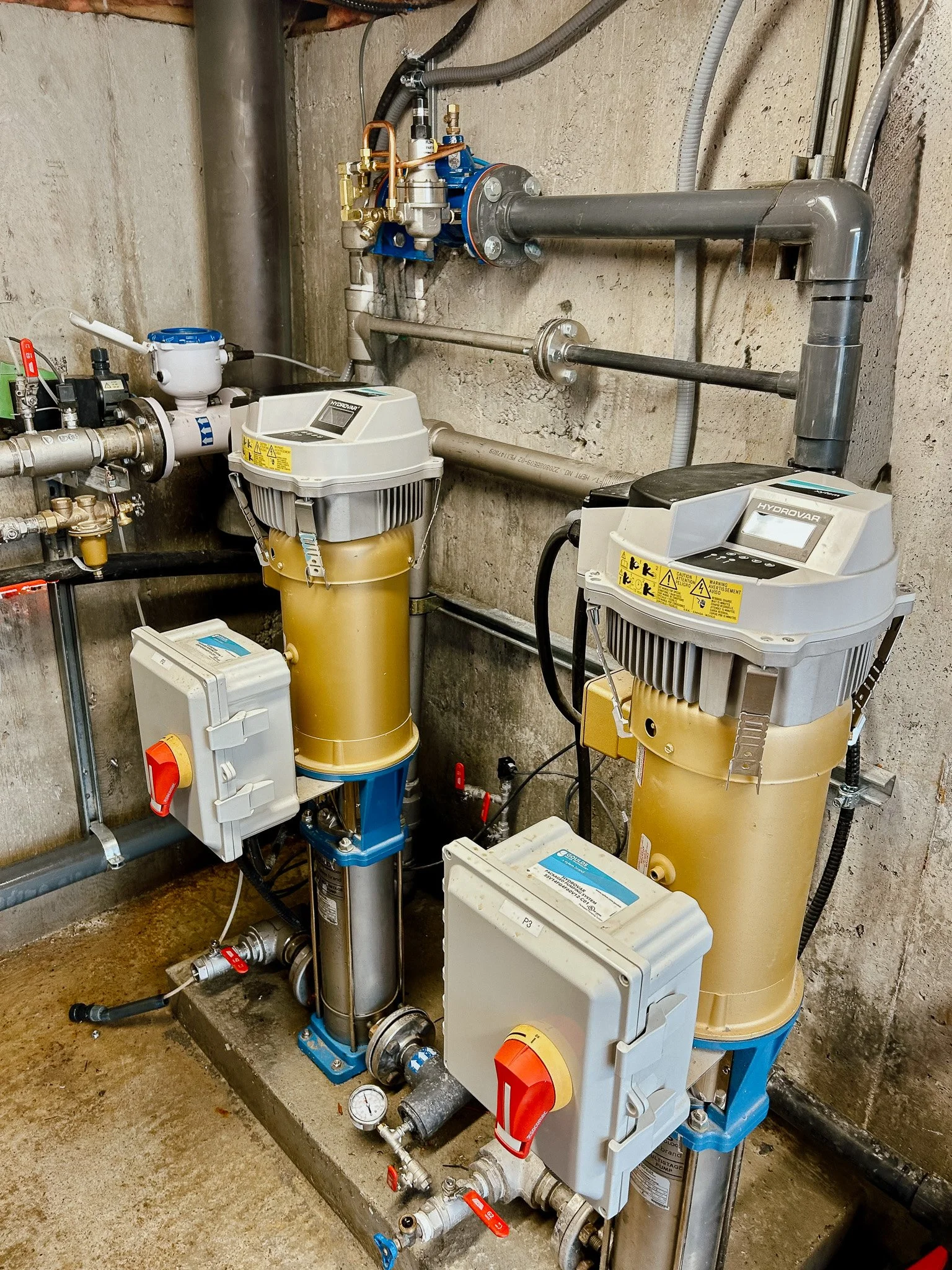 Industrial water filtration and water softening equipment with pipes and control boxes mounted on a concrete wall in a utility room.