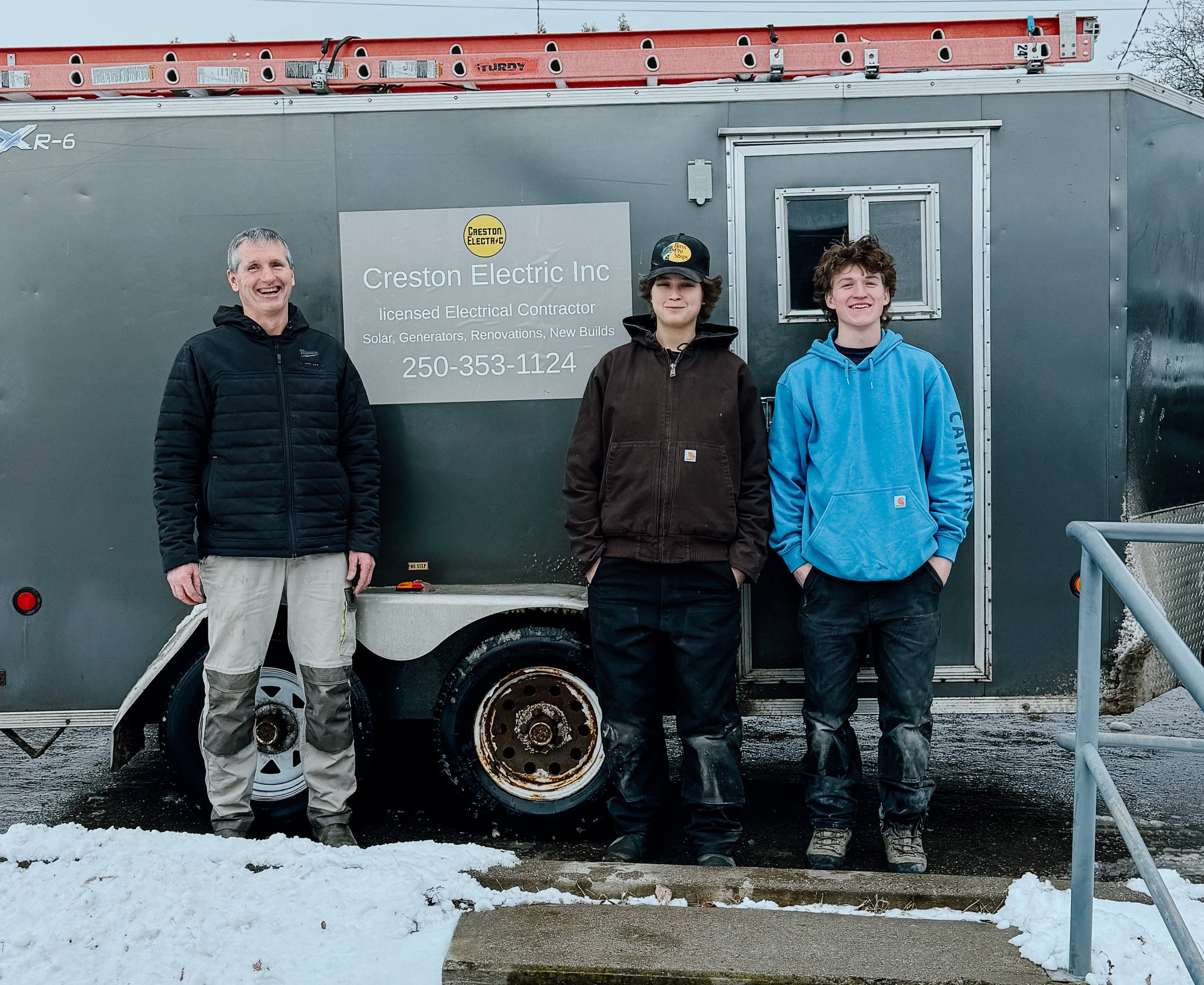 Three people standing in front of a trailer with a sign that reads 'Creston Electric Inc, licensed Electrical Contractor.