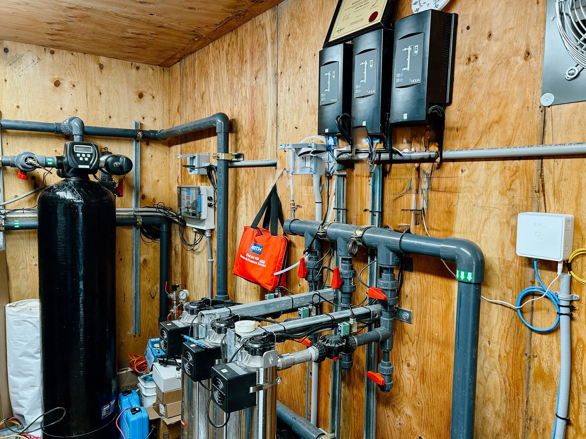 Mechanical room with water filtration and pumping equipment, including large black tank, pipes, valves, and electronic controllers mounted on a wood-paneled wall.