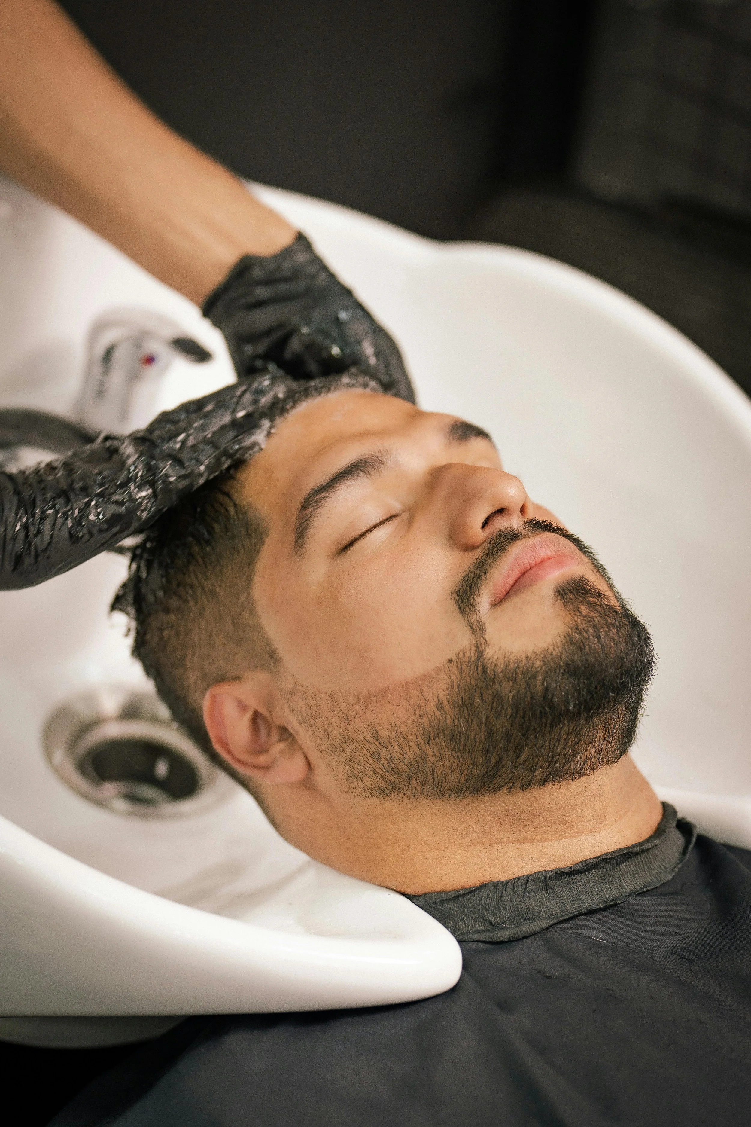 Man relaxing at a salon or barbershop while getting his hair washed in a sink with water and shampoo, with a hairstylist wearing black gloves.