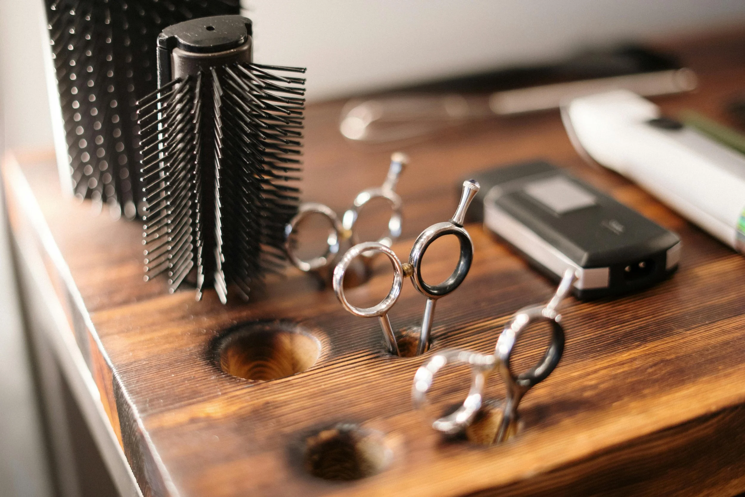 Barber tools on a wooden countertop, including a black hairbrush, scissors, a straight razor, and a clipper.