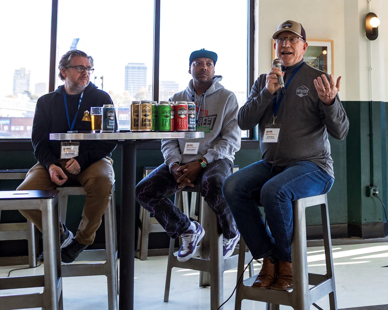 three presenters sit at a table as part of a panel discussion. one person is holding a microphone and gesturing as he speaks.