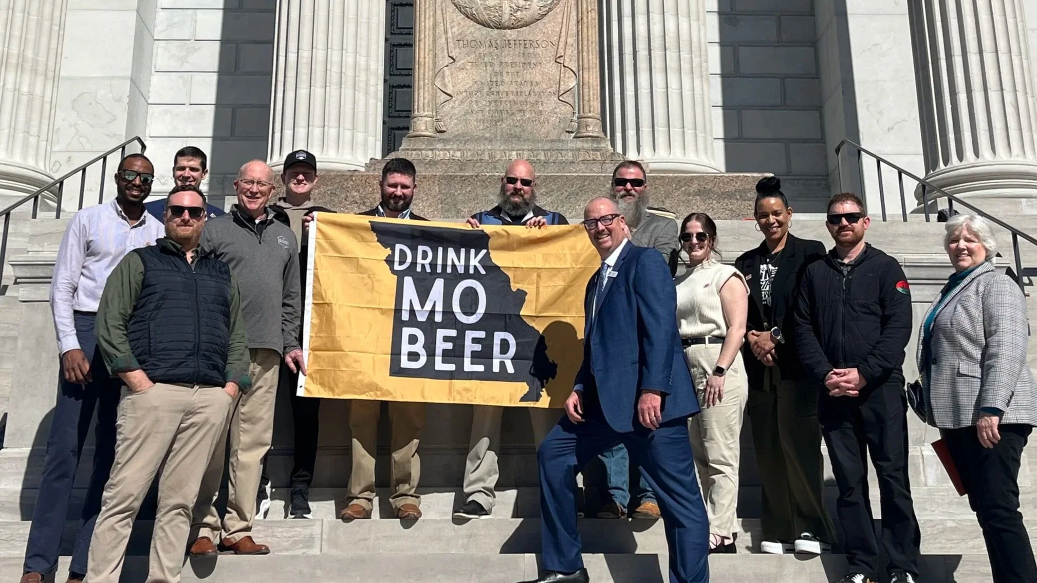 group of women and men standing outside on the steps of the missouri state capitol building holding a banner that reads drink mo beer with an icon in the shape of the state of missouri on it