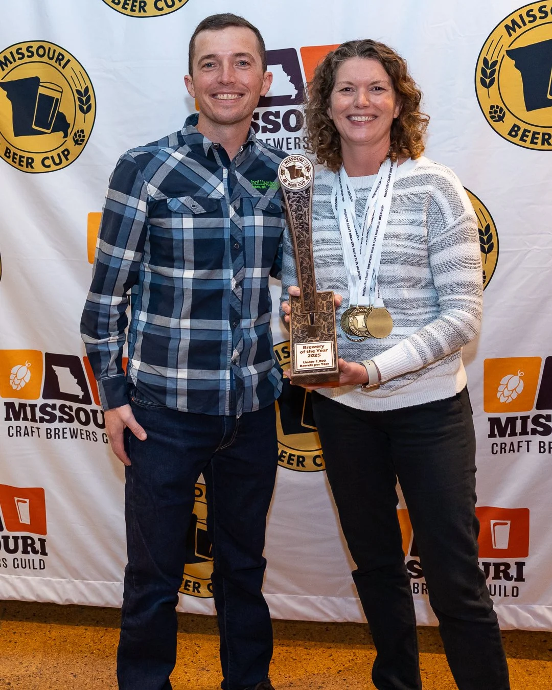a sponsor poses with a medal winner wearing several medals and holding the brewery of the year trophy