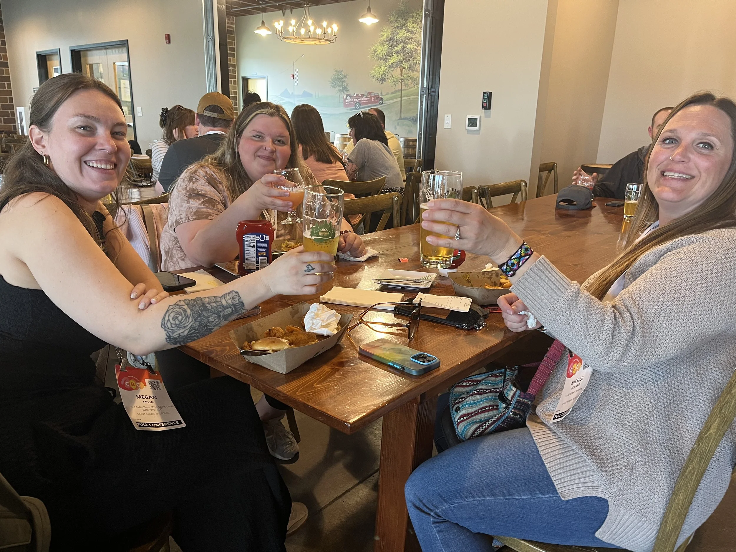 three women sit at a table and hold up their glasses in a cheers gesture