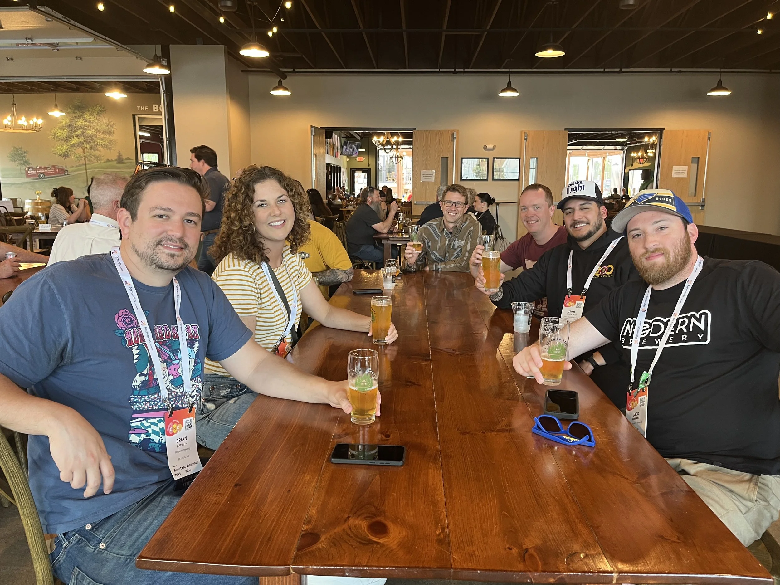 Seven people seated at a table in a brewery hold their glasses in a cheers gesture