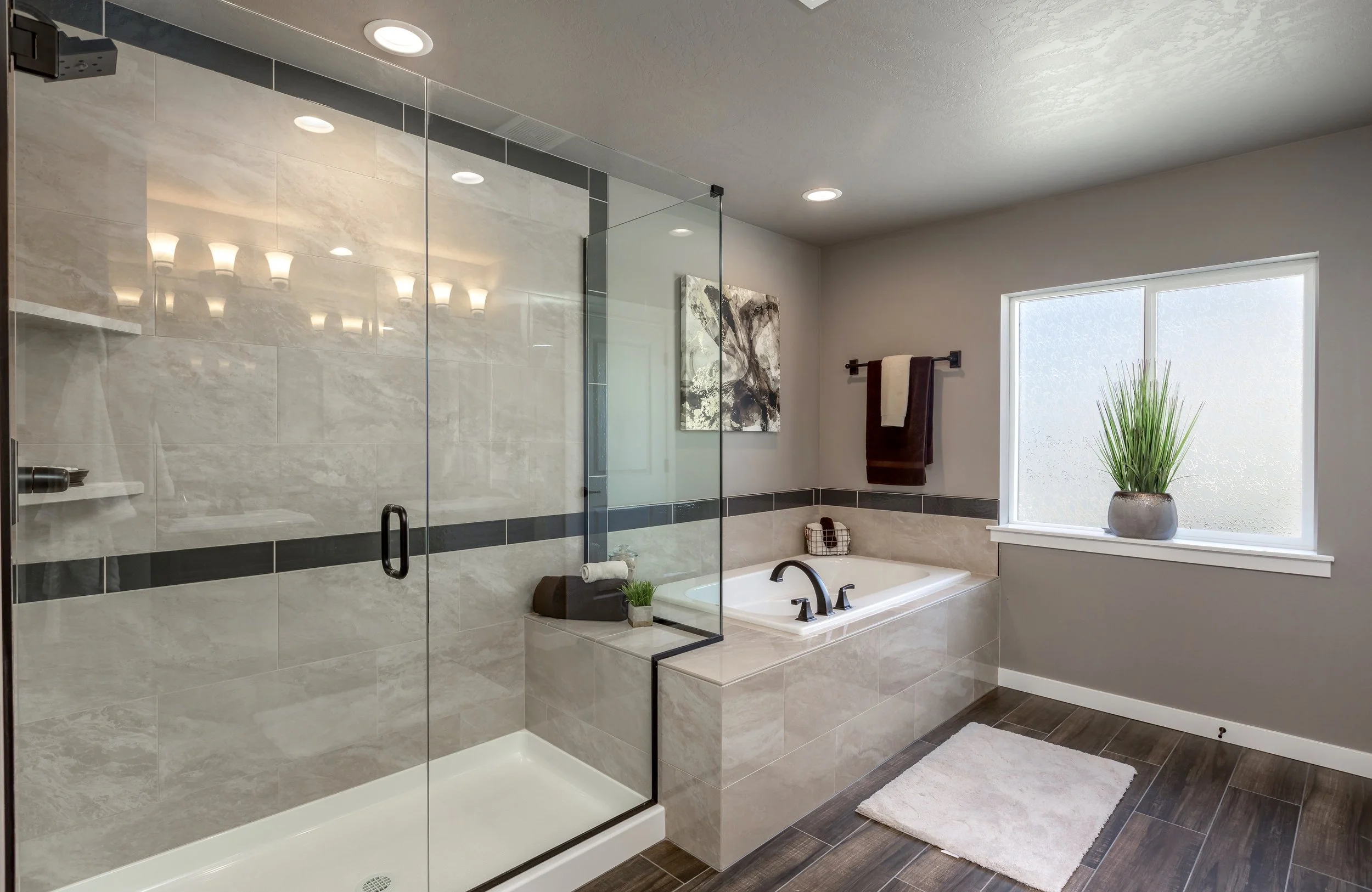 Modern bathroom with a glass shower enclosure, a bathtub with a black faucet, a large frosted window, and a potted plant on the window sill.