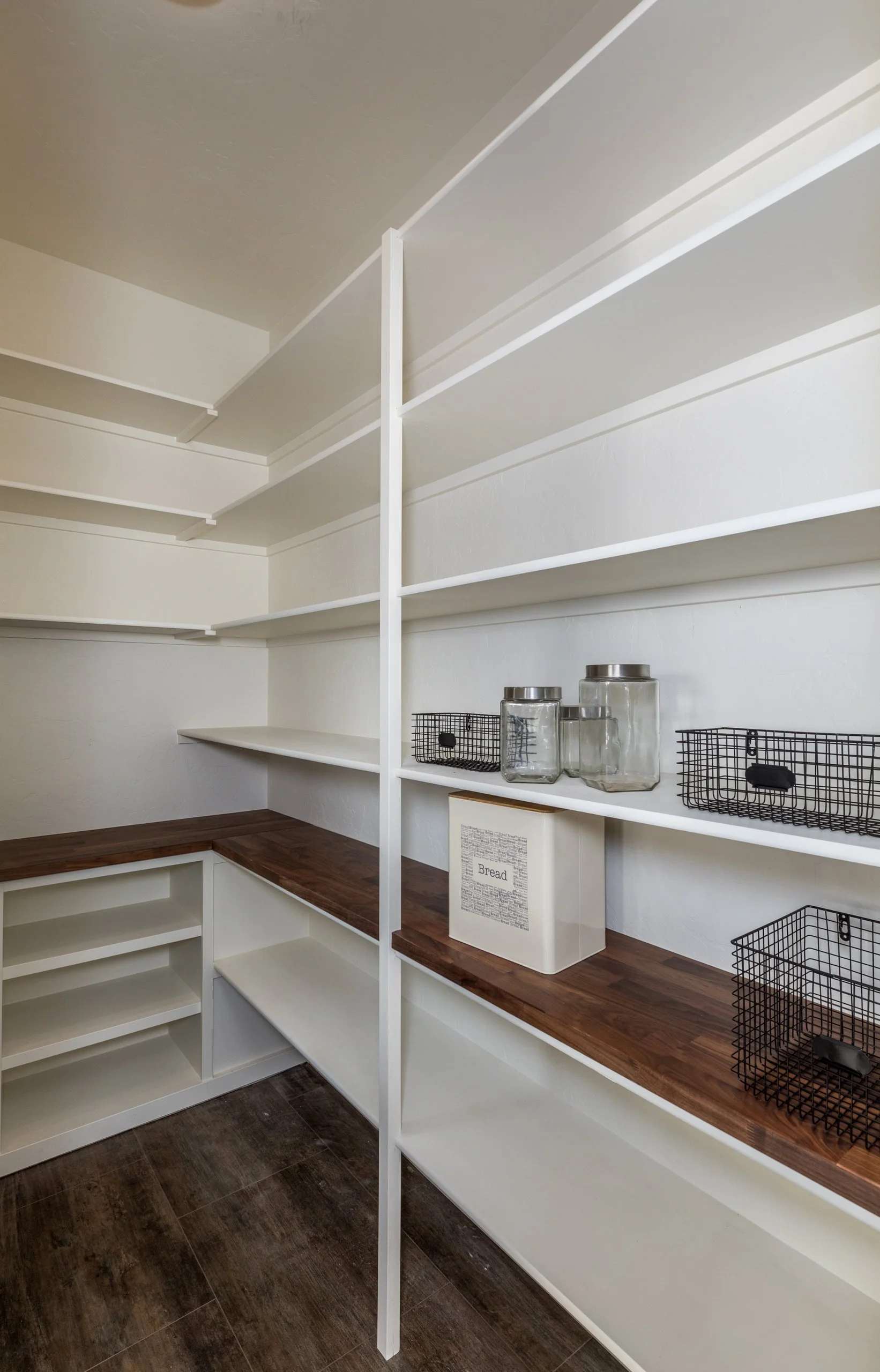 Staged simply, empty pantry with white shelves, wooden countertop, glass jars, and wire baskets.