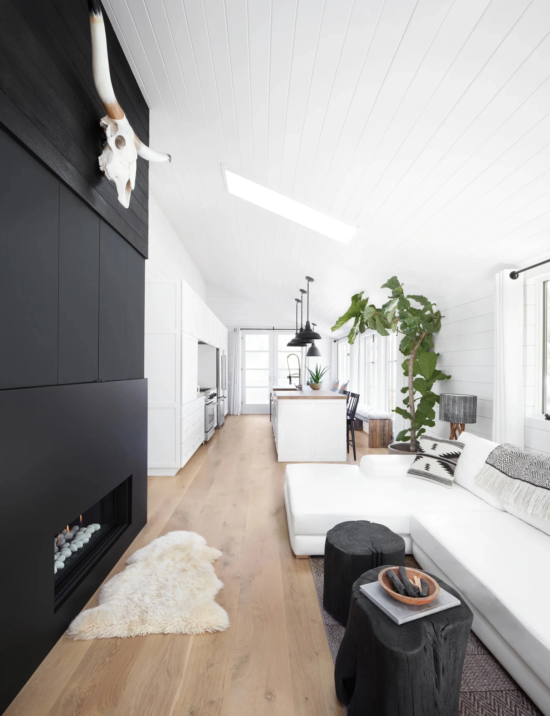 Bright modern living room with white walls, wood floors, black fireplace, white sectional sofa, decorative pillows, tall houseplant, black side tables, kitchen area with island, pendant lights, and large windows.