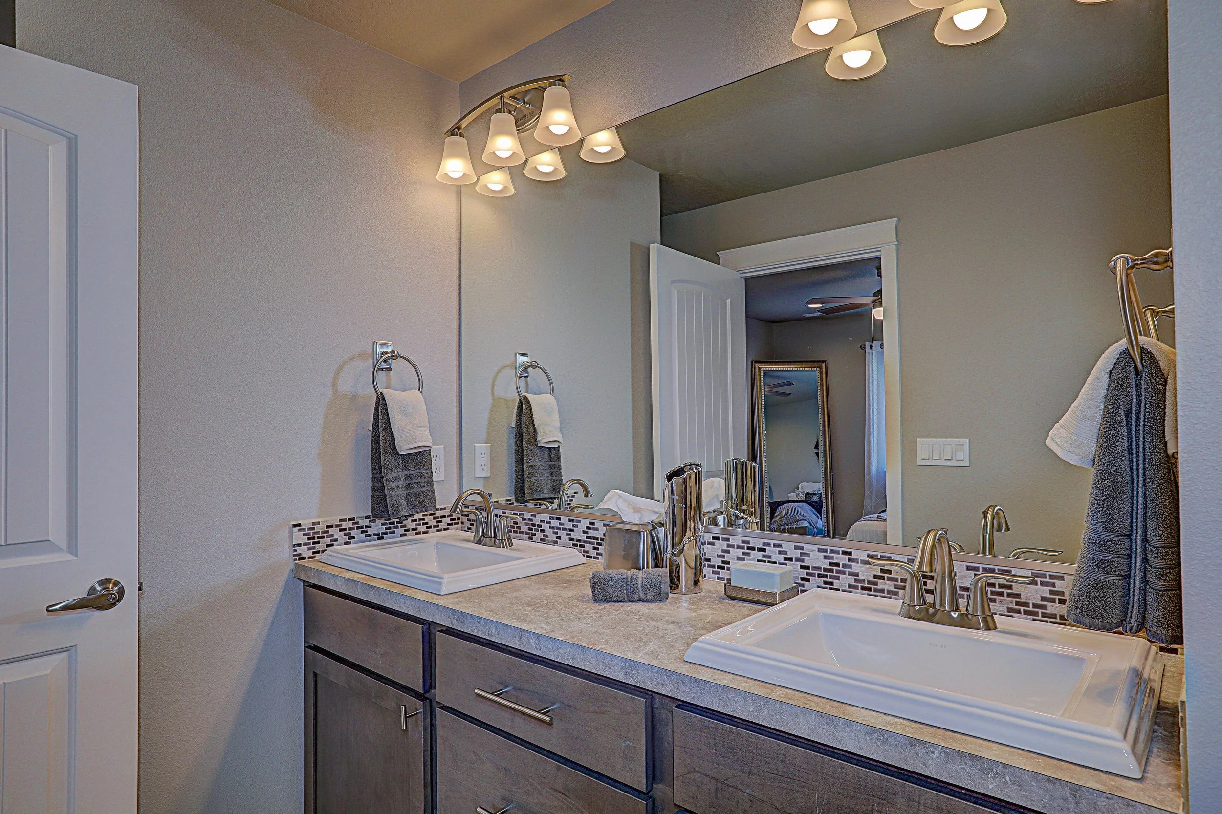 A modern bathroom vanity with dual sinks, a large mirror, and a set of lighting fixtures overhead. Towel rings with towels are mounted on the wall, and the vanity has dark wood cabinets.