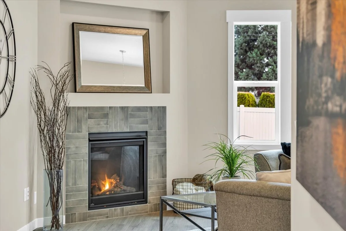 Living room with a fireplace, a large mirror above it, a window with a view of trees and a fence, and a beige sofa with a green plant nearby.